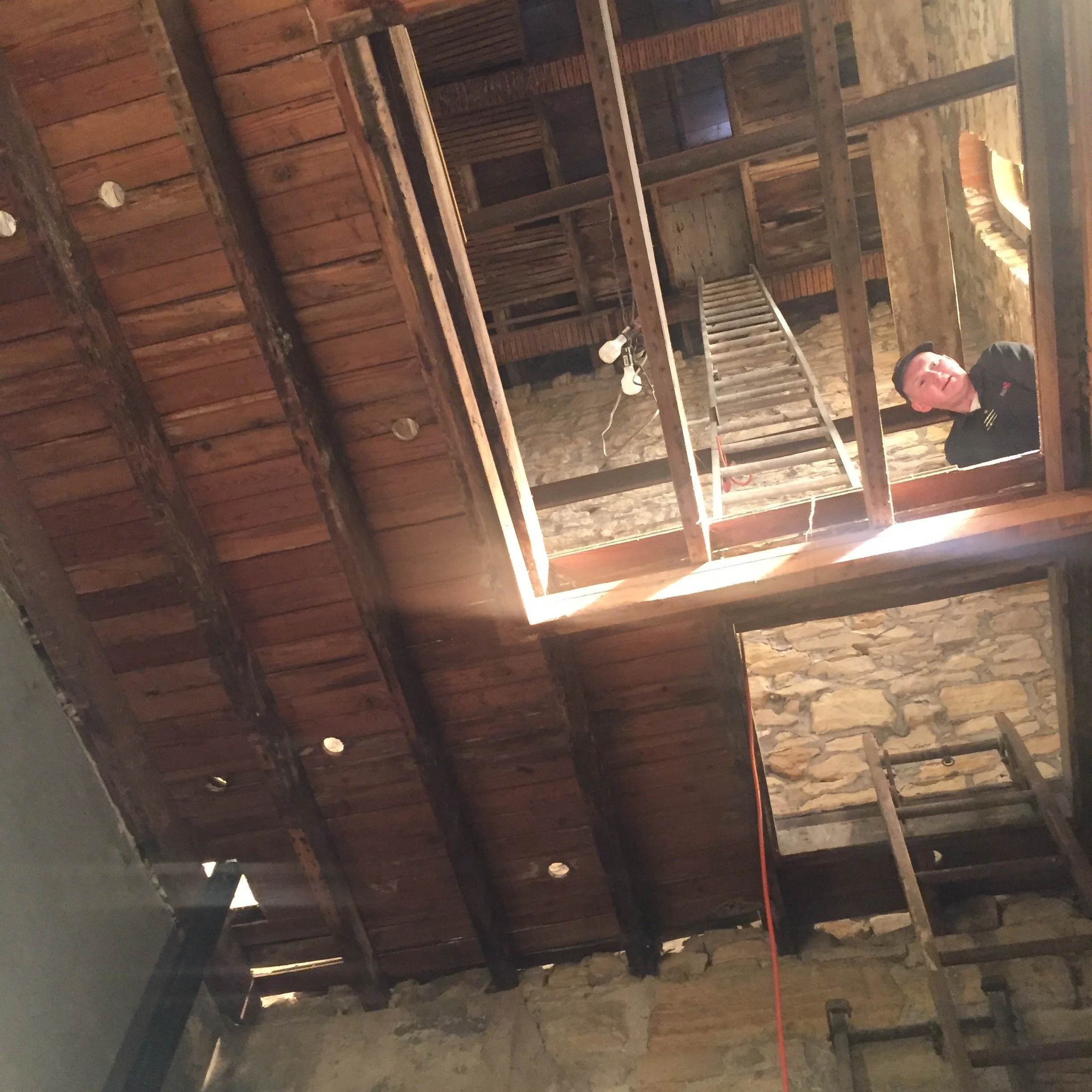 Interior of a building under construction with exposed wooden beams, brickwork, a ladder, and construction light. Part of a person wearing a dark shirt and cap is visible in the bottom right corner.