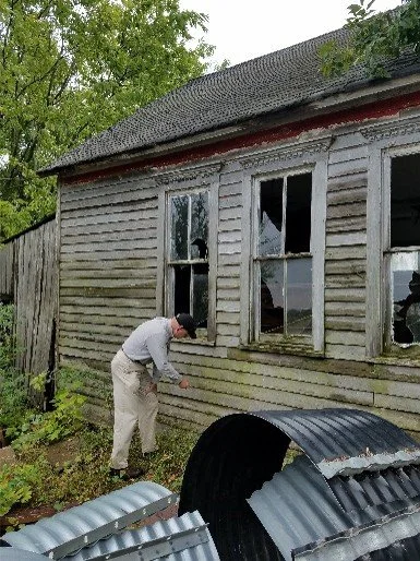 An architect examining the weathered exterior wall of an old wooden house with multiple broken windows.