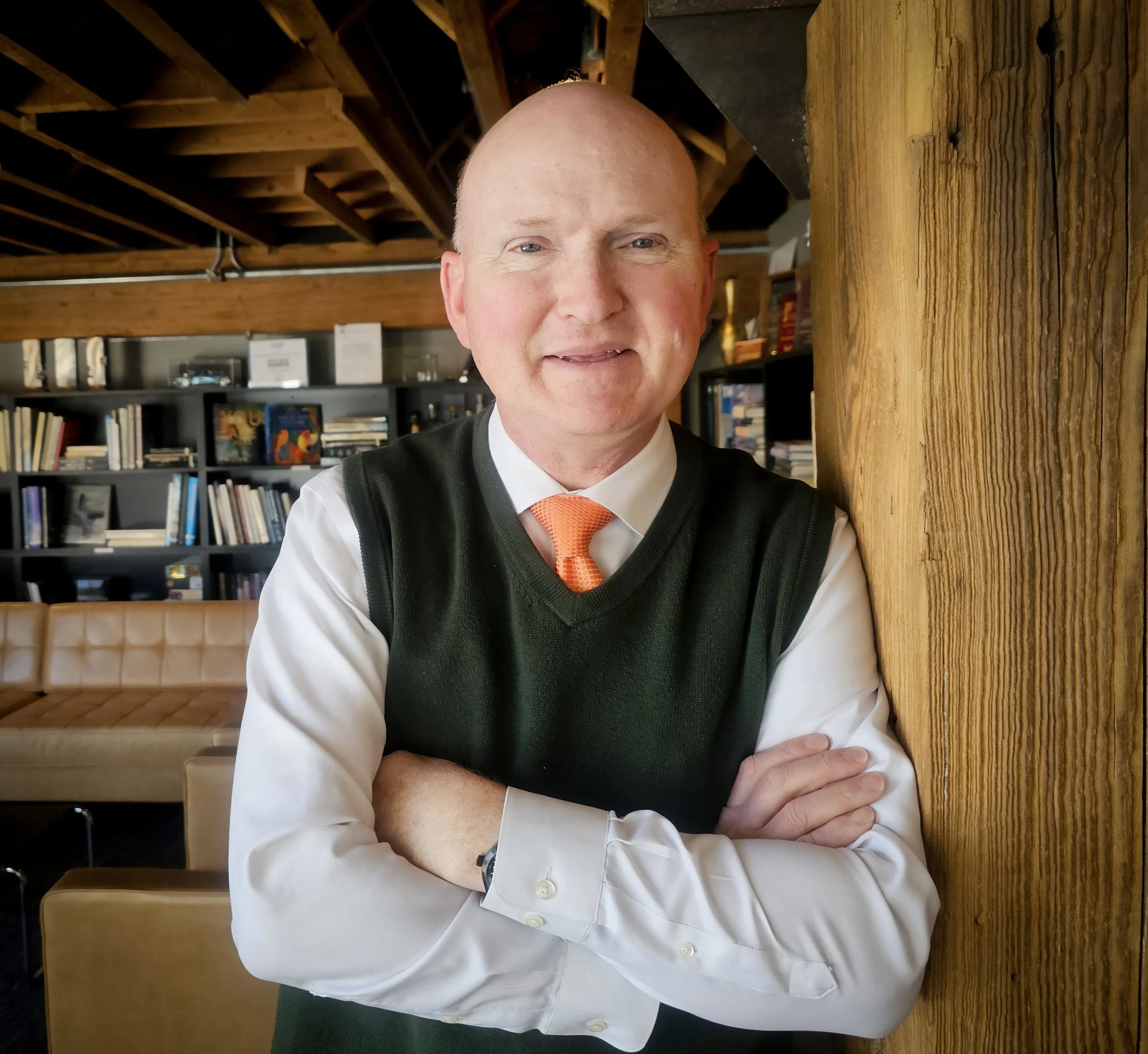 A man with a bald head, light skin, wearing a white shirt, a dark sweater vest, and an orange tie, in front of a bookshelf filled with books and decorative items in a cozy, wood-paneled room.