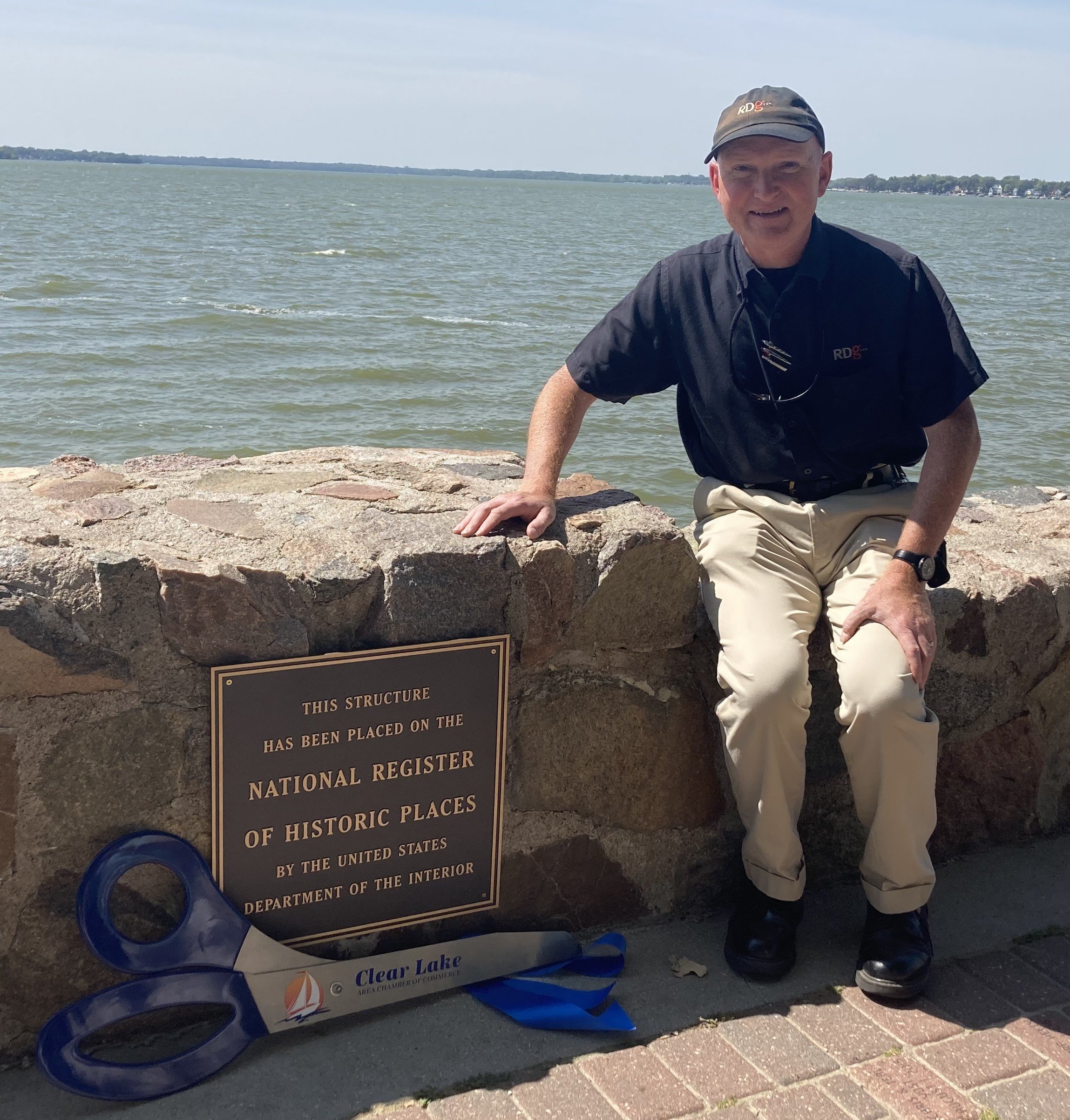 A man sitting on a stone ledge near a large body of water, wearing a black shirt and beige pants, with sunglasses hanging from his shirt collar. There is a plaque on the stone structure and a blue paddle with a logo resting on the ground next to him.