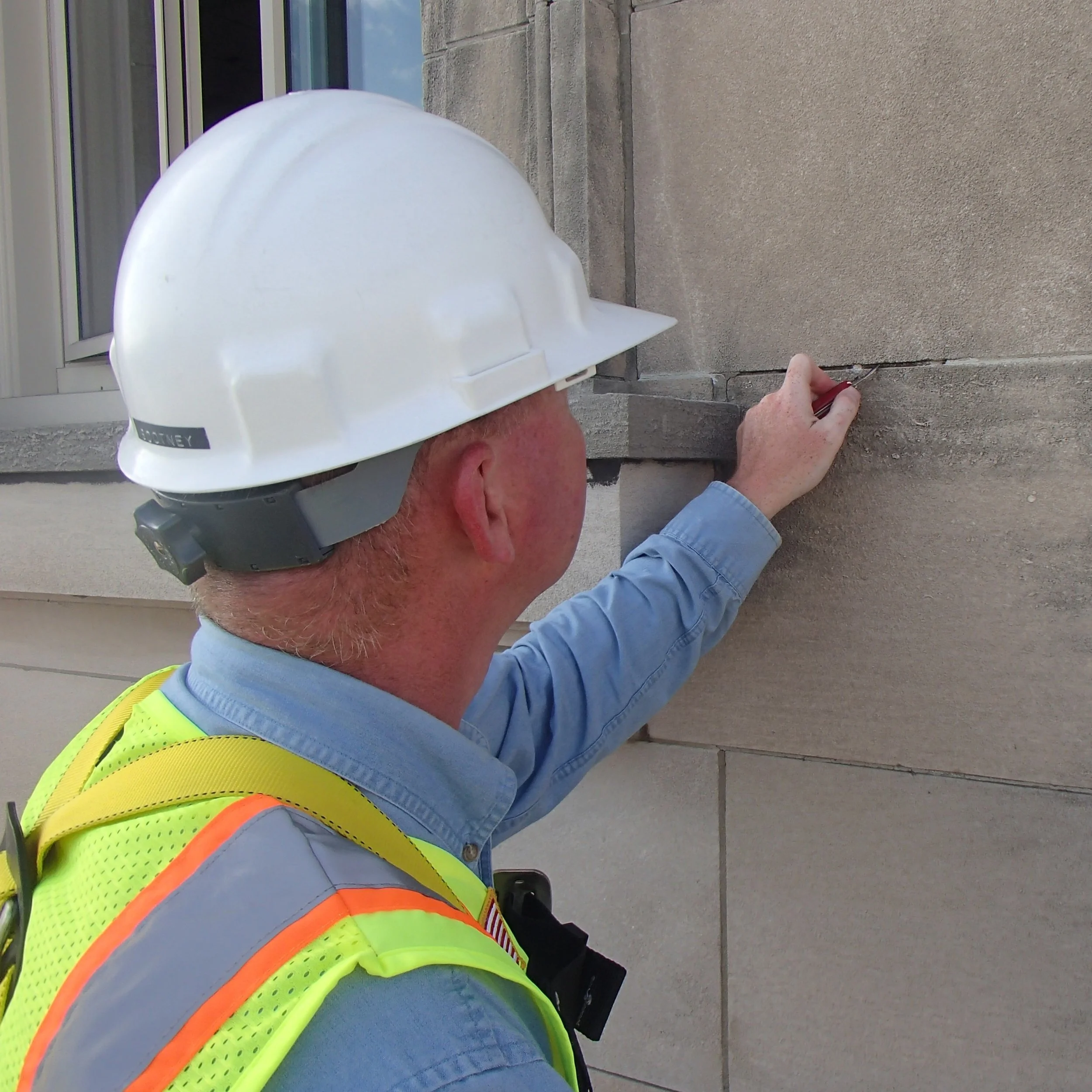 An architect wearing a white hard hat and a neon safety vest is examining stone masonry on a building wall outside.