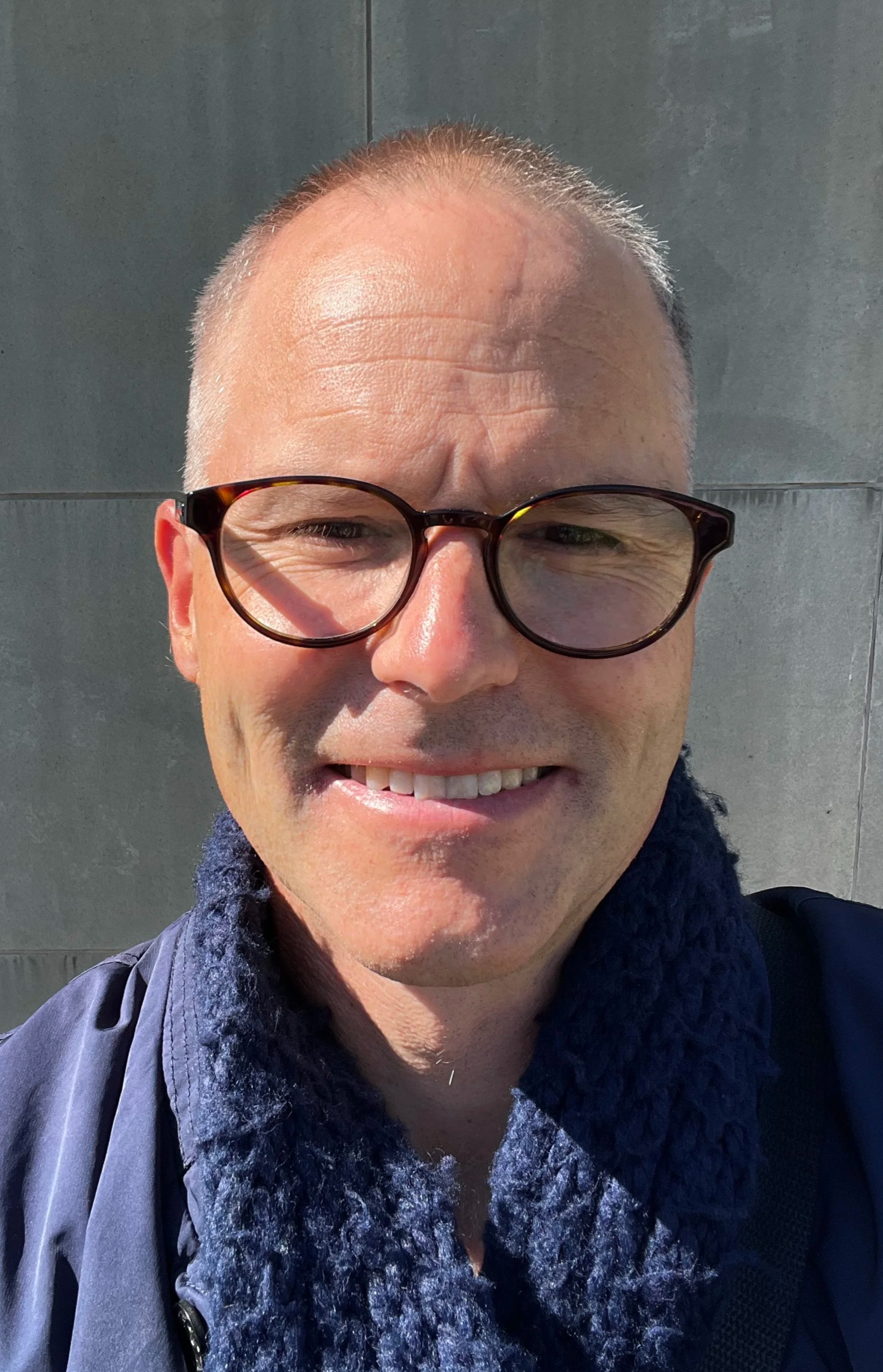 Close-up of a man wearing glasses, smiling outdoors on a sunny day. He has a buzz cut hairstyle, light skin, and is dressed in a dark jacket with a fuzzy collar.