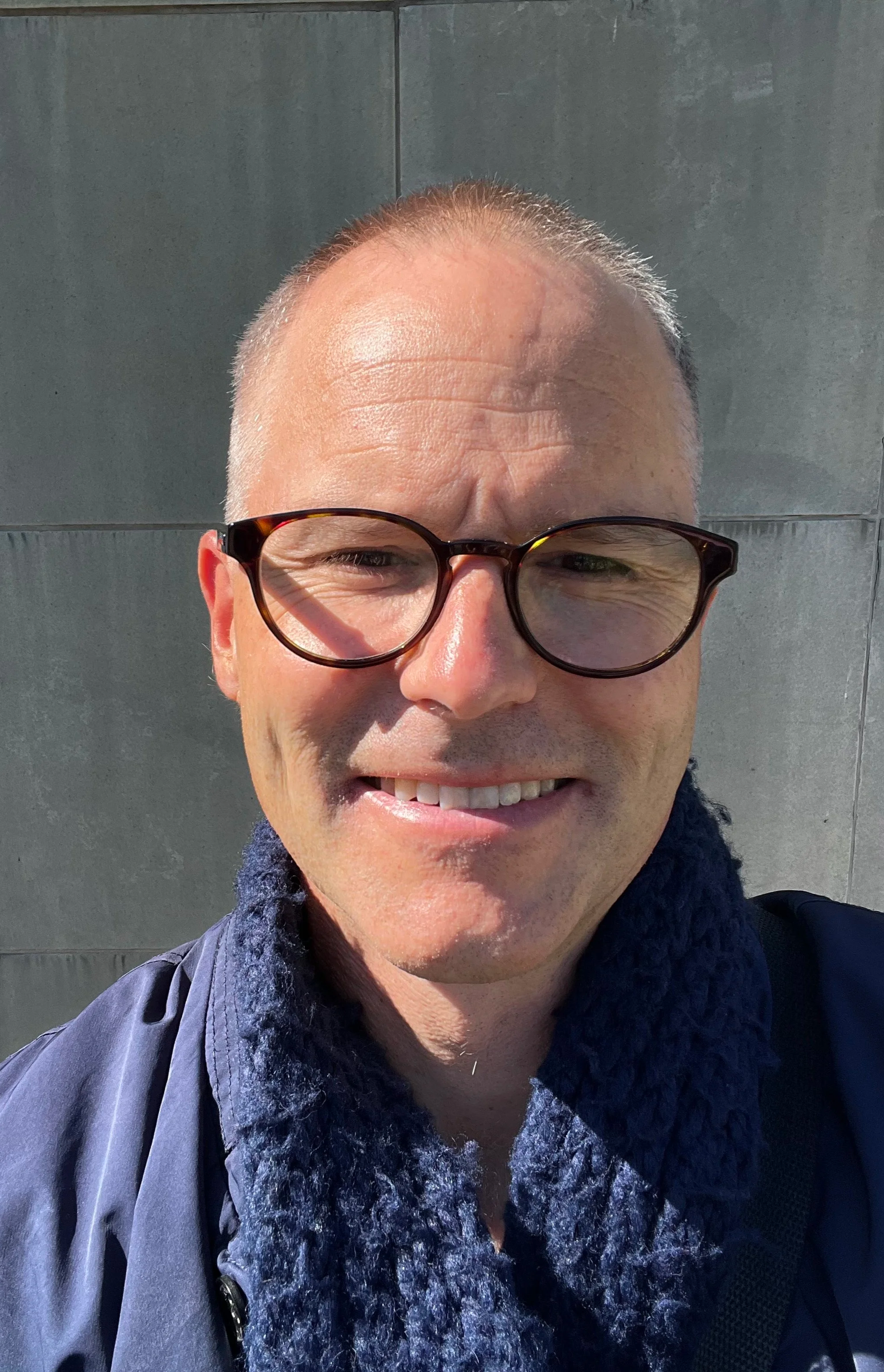 A man smiling outdoors, wearing glasses and a dark jacket with a fuzzy scarf, standing against a wall.
