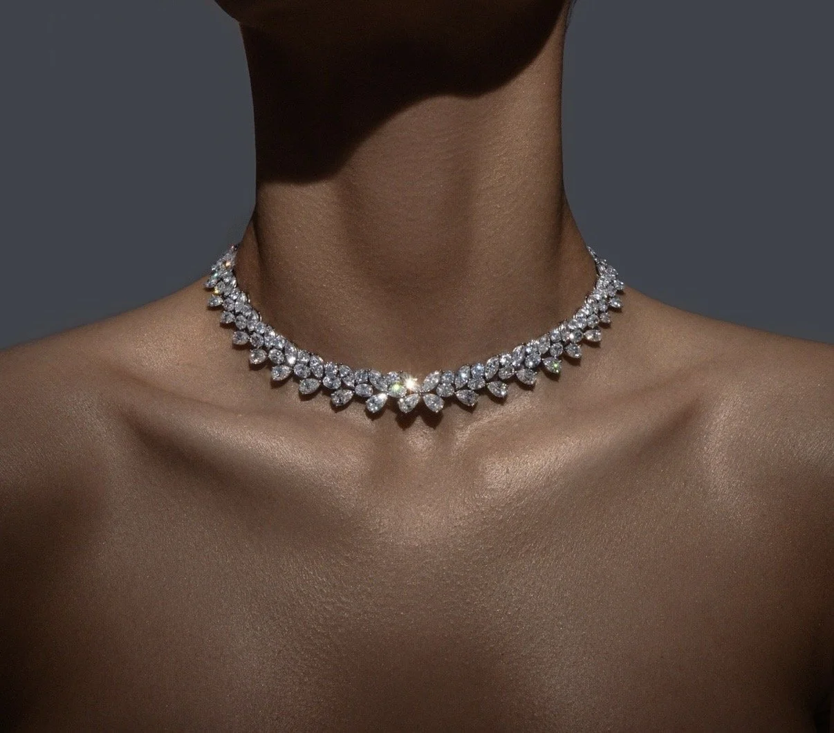 Close-up of a woman's neck and shoulders with a diamond necklace against a neutral background.