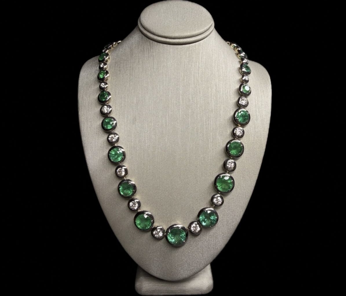 Necklace with alternating green gemstones and clear diamonds on a white display bust against a black background.