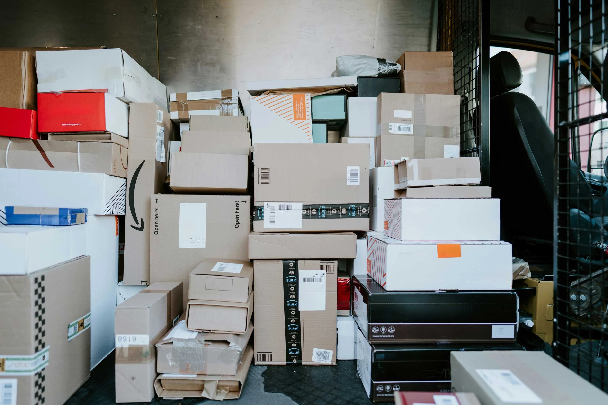 Stacked cardboard boxes and packages in a warehouse or delivery vehicle interior, with black metal shelving to the side and a car seat visible in the background.