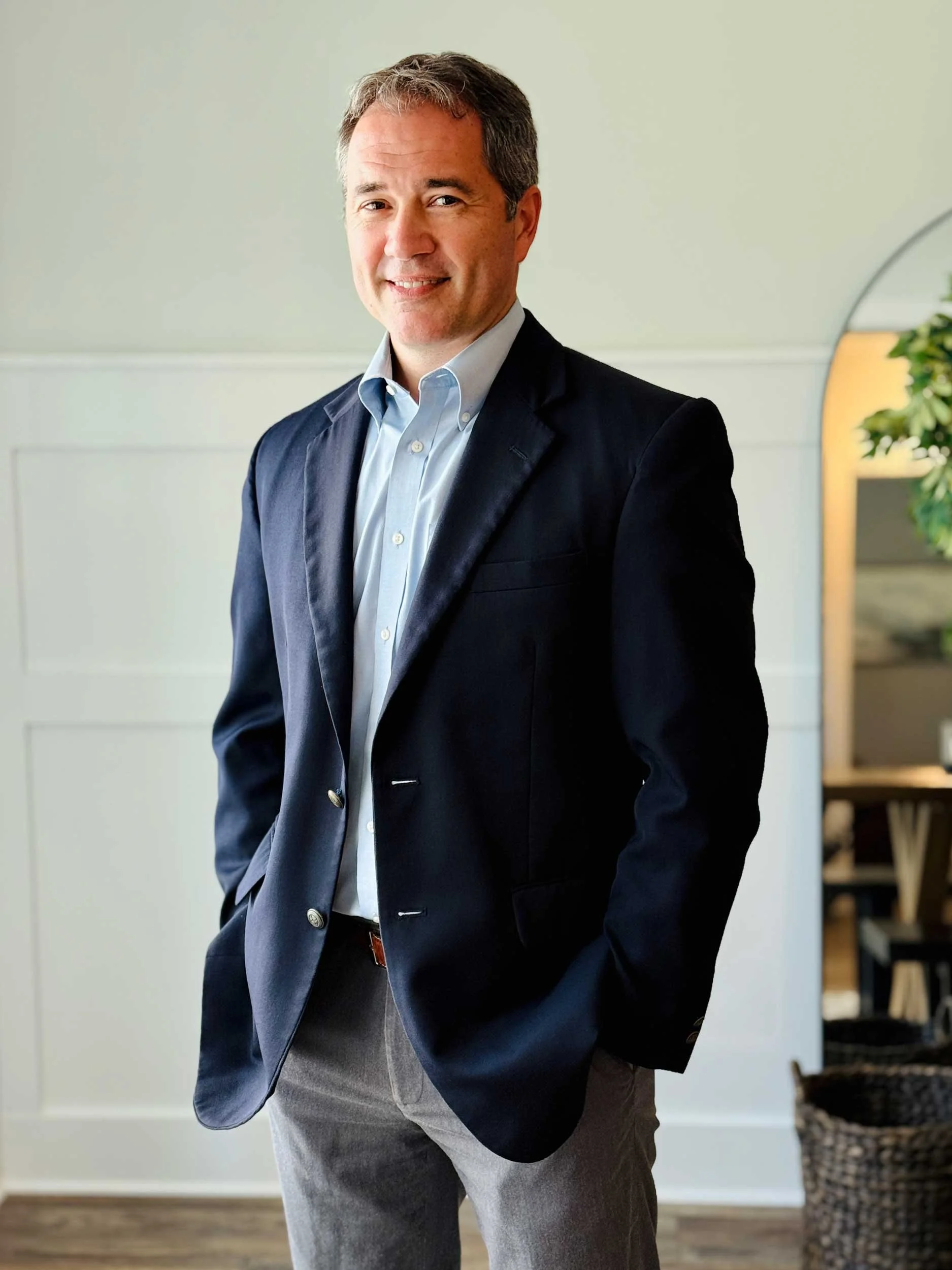A man wearing a dark blazer, light blue shirt, and grey pants, standing indoors with a neutral expression, in a room with light-colored walls, wooden flooring, and a wicker basket in the background.