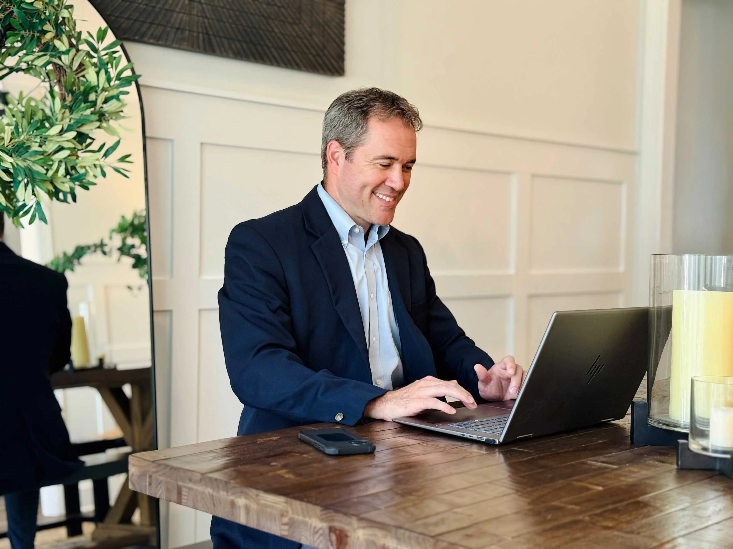 A man in a suit sitting at a wooden table working on a laptop in a bright, modern room.