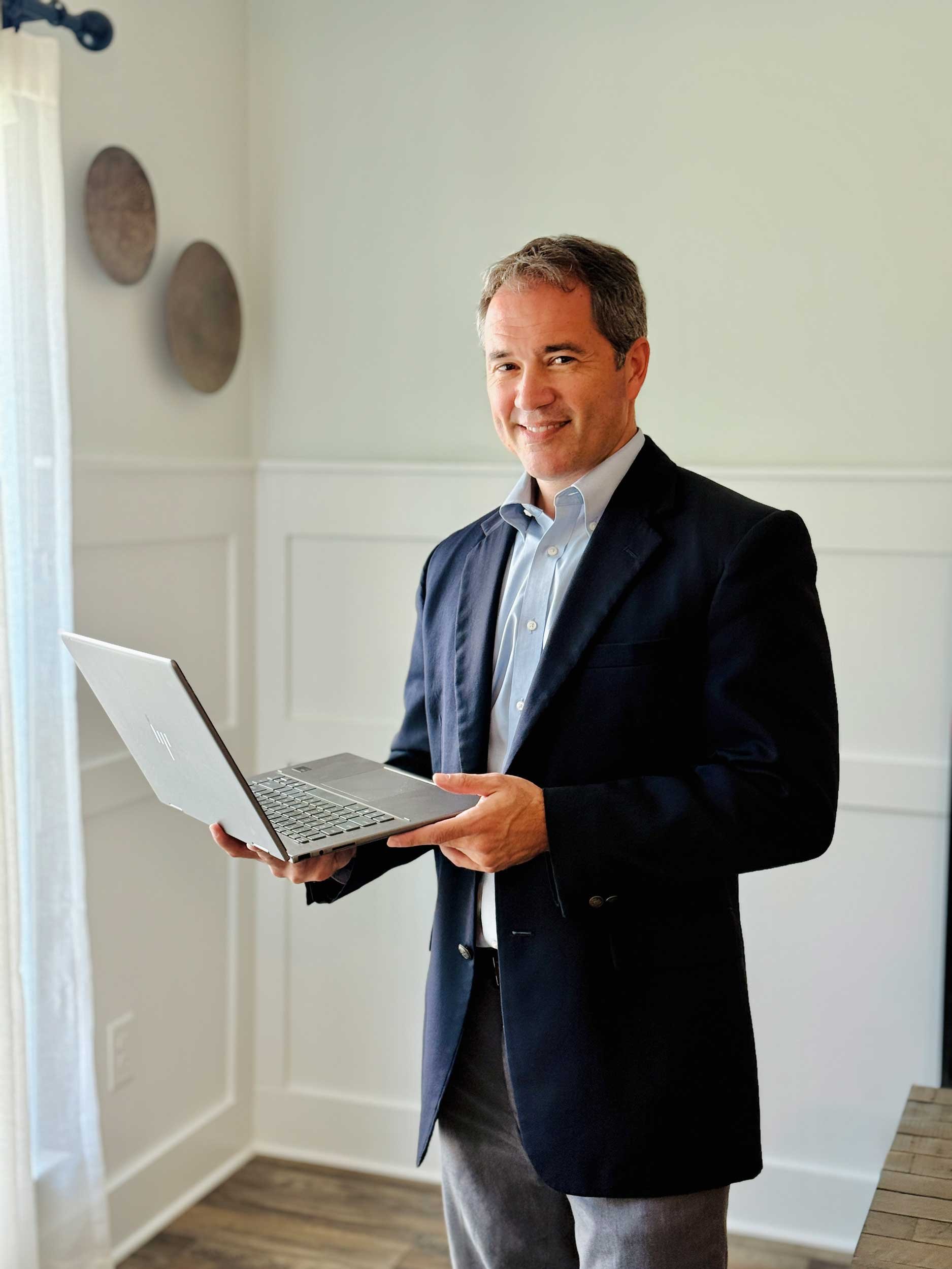 A man in a dark suit and light blue shirt holding a silver laptop in a room with white walls, hardwood floors, and decorative wall plates.
