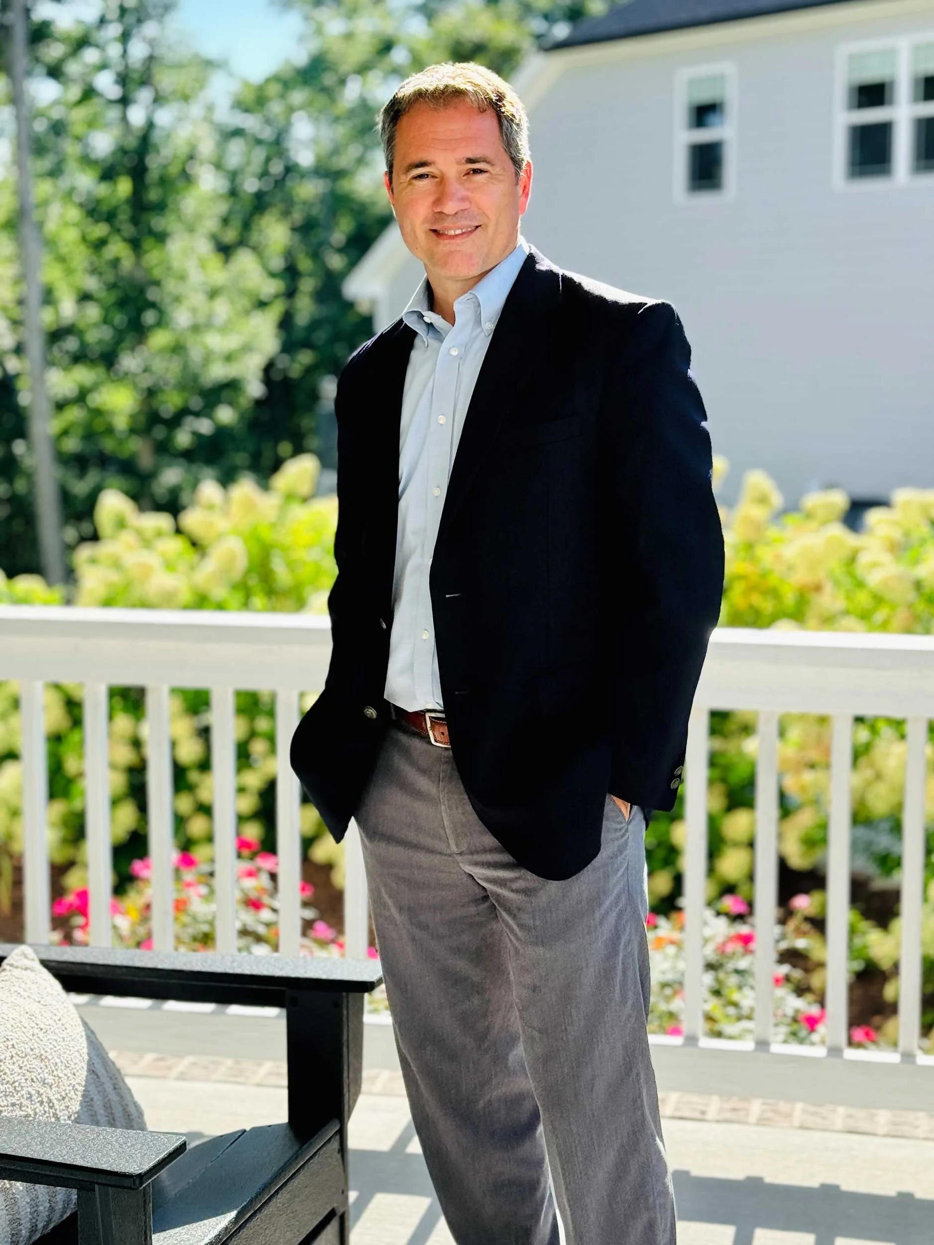 A smiling man with short brown hair, wearing a white shirt and black blazer, standing outdoors in front of a white house with multiple windows and surrounded by green trees and colorful flowers.