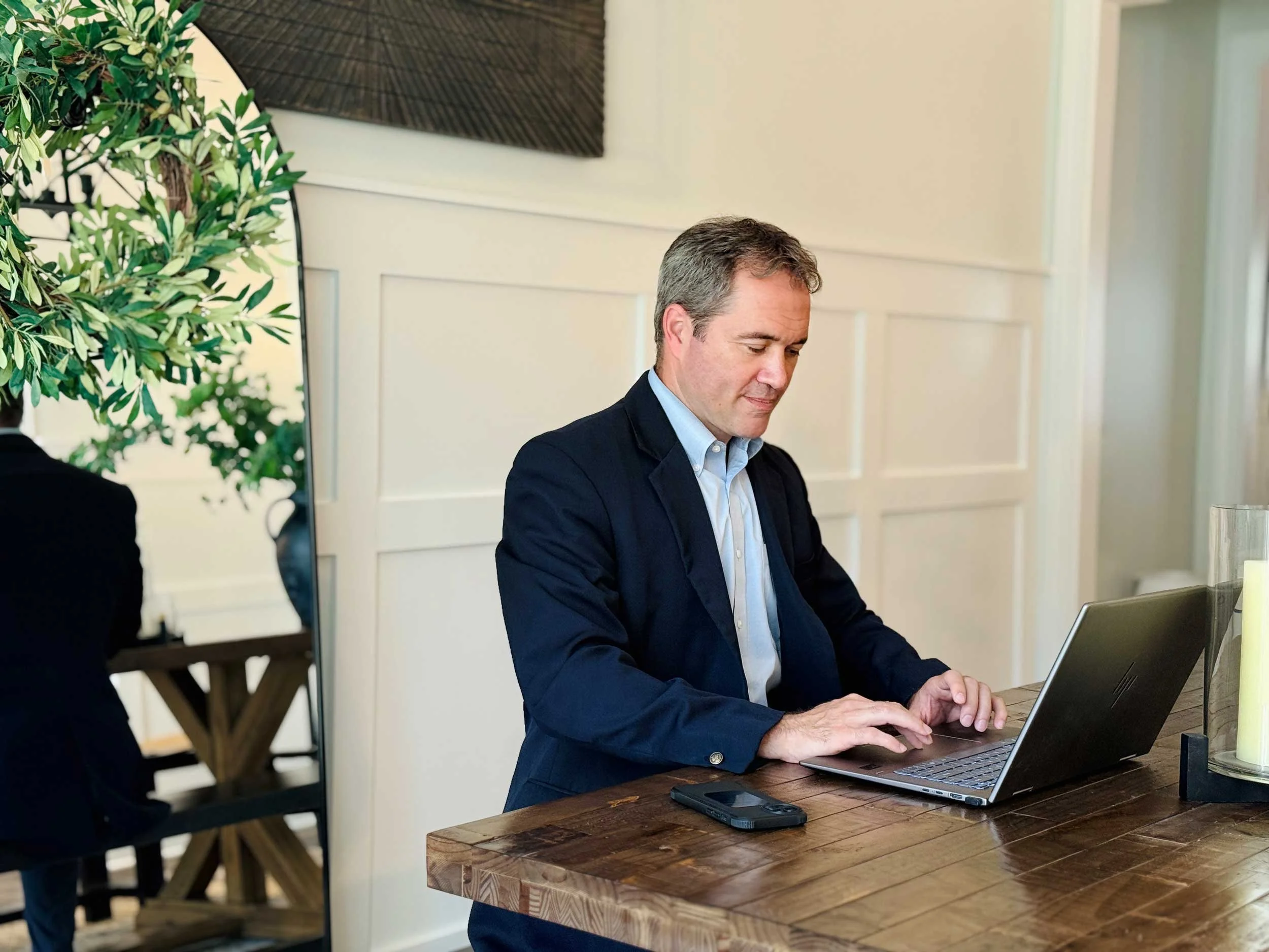 A man in a blue blazer and light blue shirt sitting at a wooden table with a laptop open in front of him, working or browsing. There is a large green plant and a framed piece of wall art behind him.