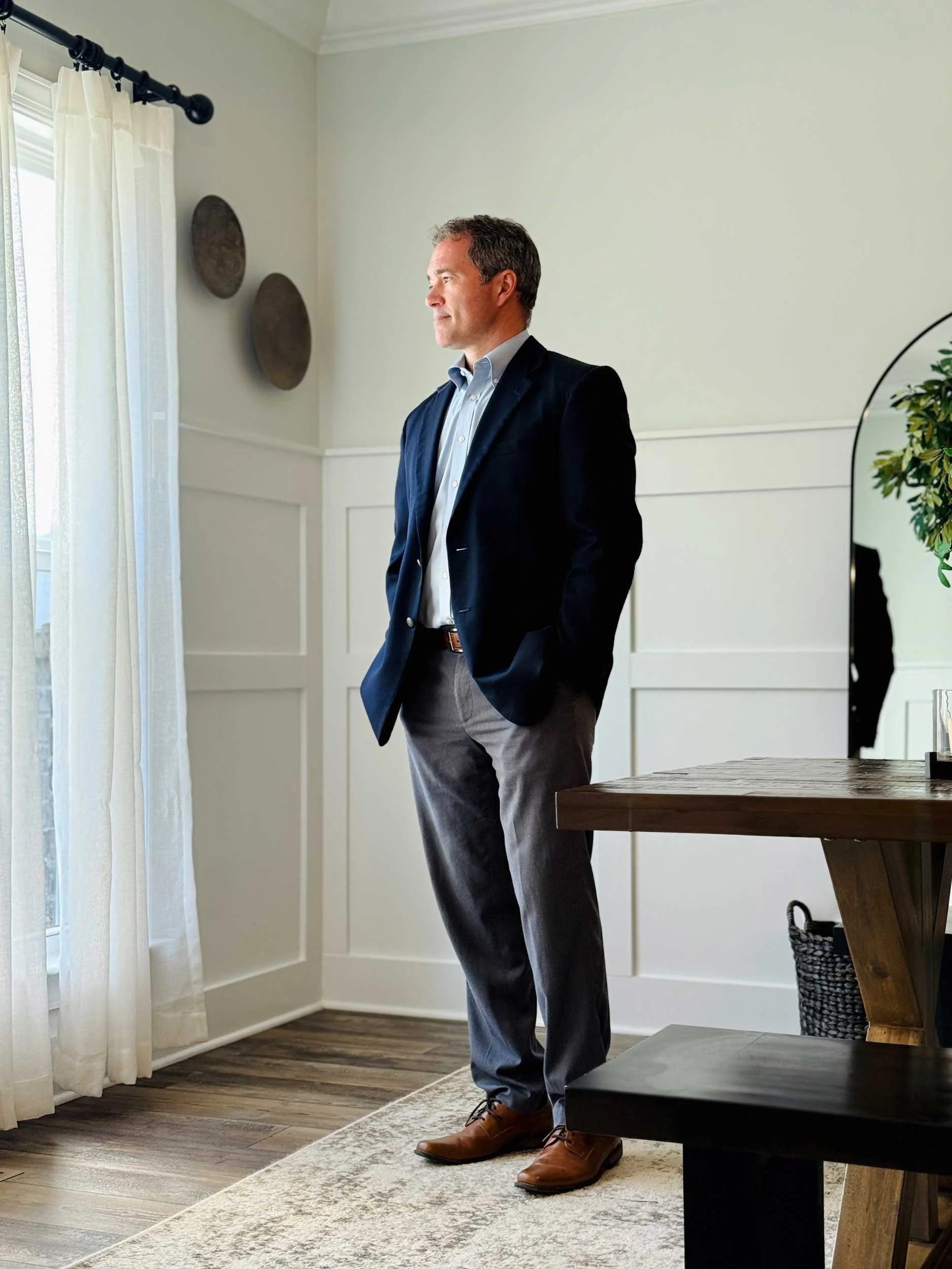 A man in business attire, wearing a dark blazer and gray pants, stands in a room with light-colored walls, looking out the window. The room has wooden flooring, white trim, cream curtains, wall art, and a wooden table.