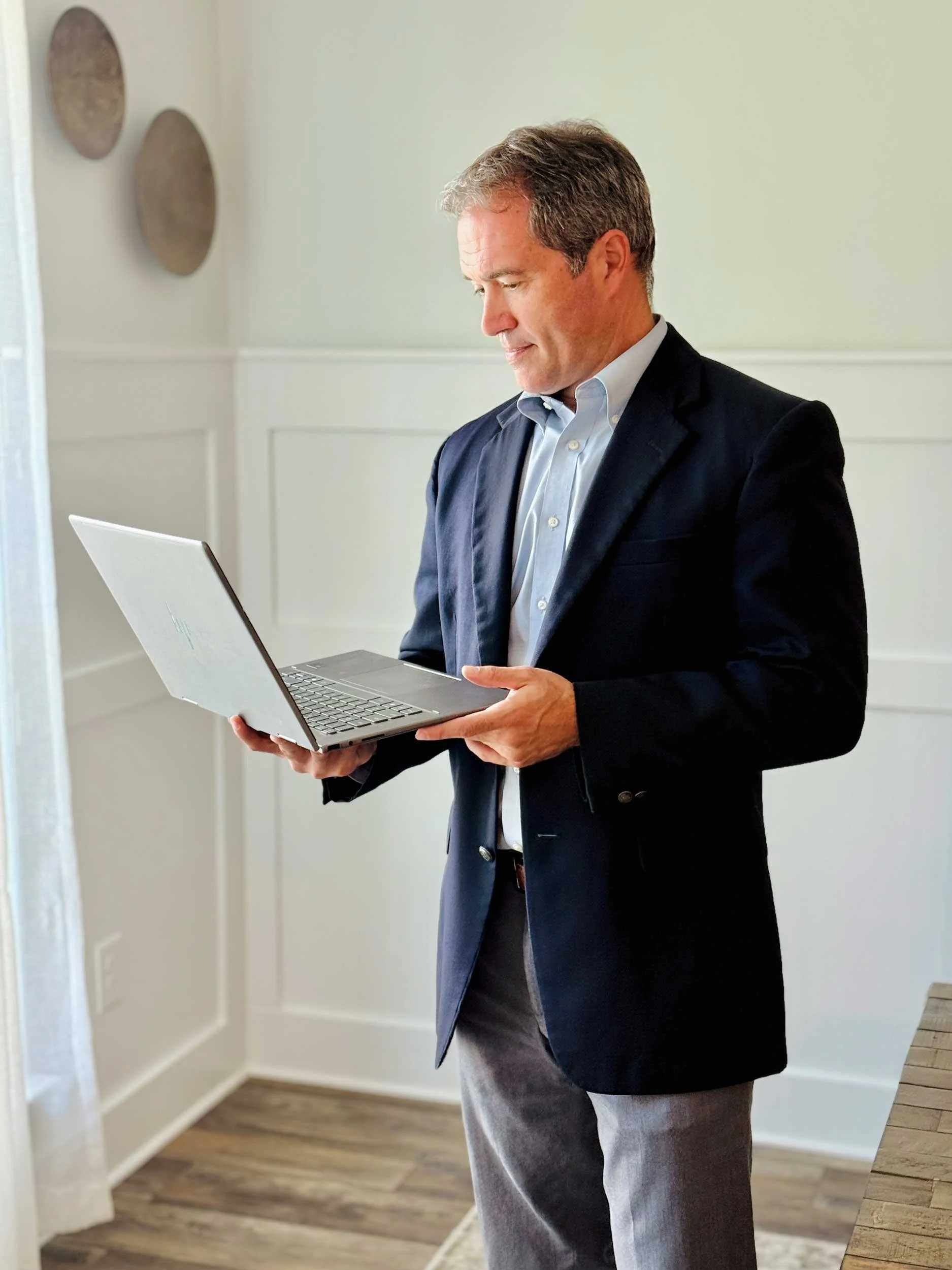 A man in a dark suit jacket, light blue shirt, and gray pants is standing in a room, looking at a laptop he is holding with both hands.