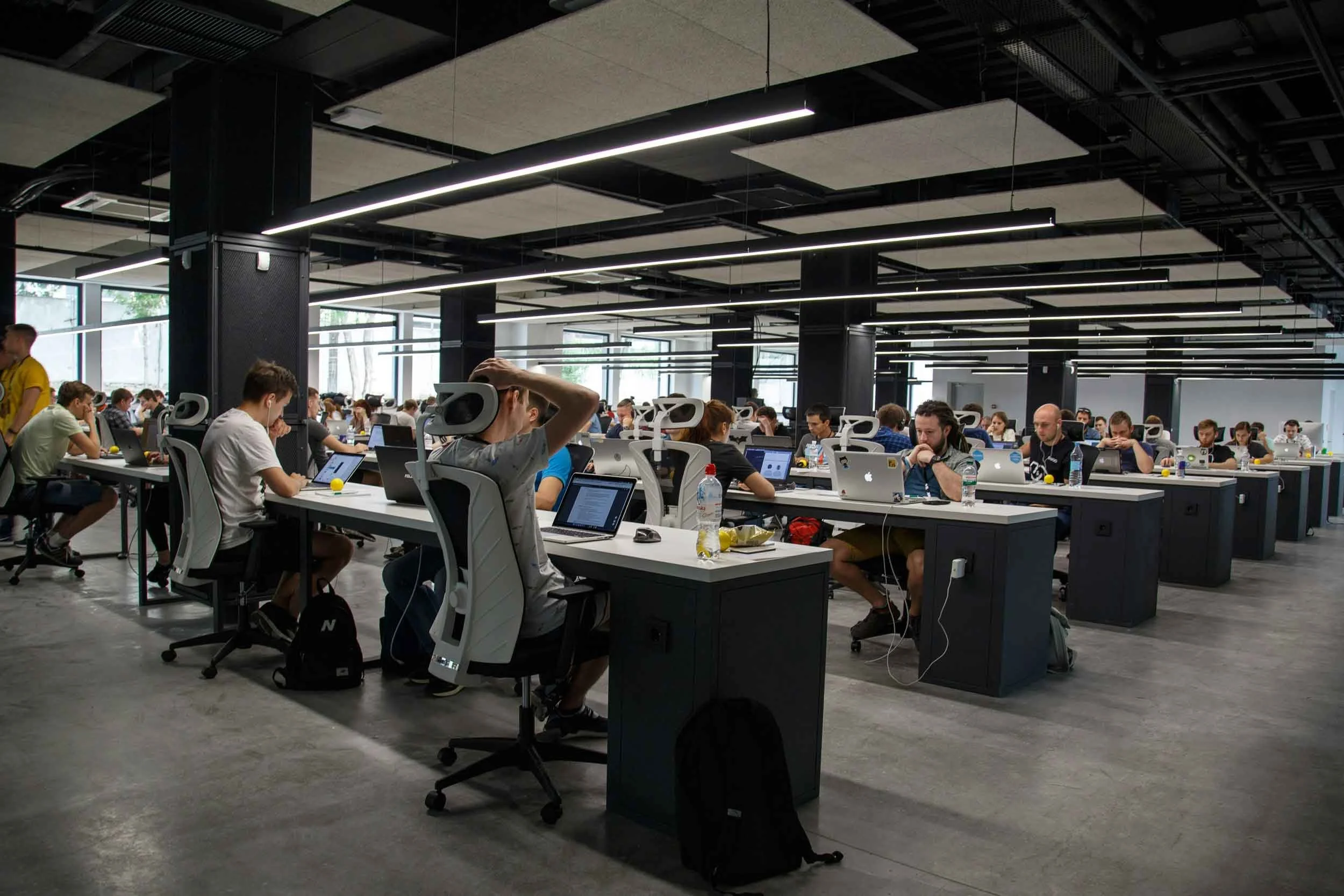 A large open-plan office filled with people working at desks with laptops. The office has modern lighting and large windows.