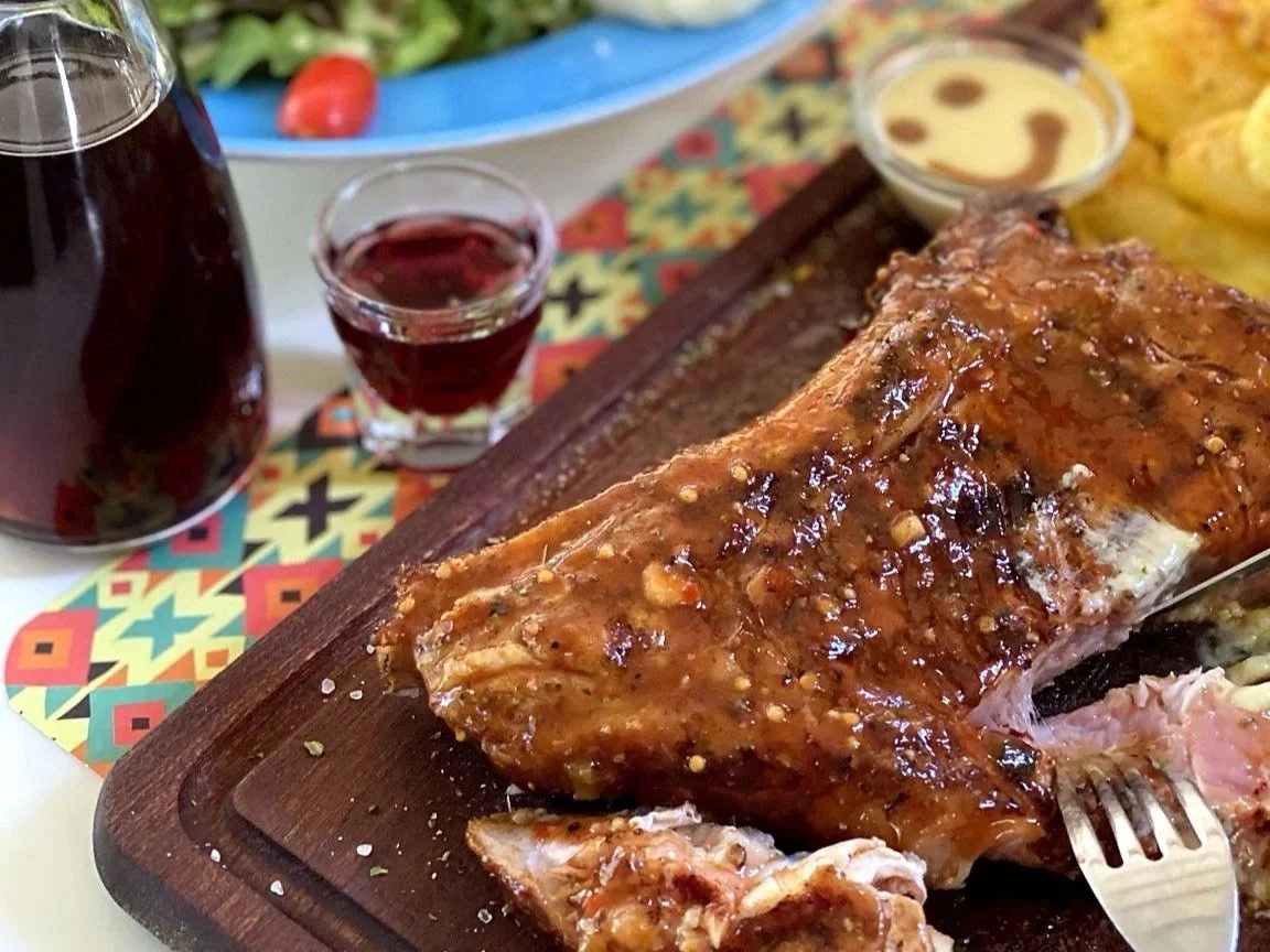 Close-up of roasted meat with sauce on a wooden cutting board, with side dishes including chips, condiments, and beverages in the background.