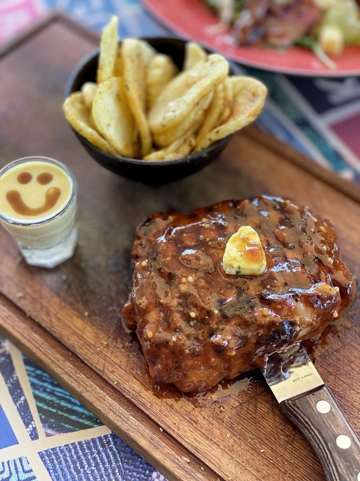 Kozis Rib Eye steak. A serving of grilled meat topped with sauce and butter, a side of French fries in a black bowl, and a shot glass with a smiley face made of sauce or ketchup on top of a creamy white beverage, all on a wooden tray.