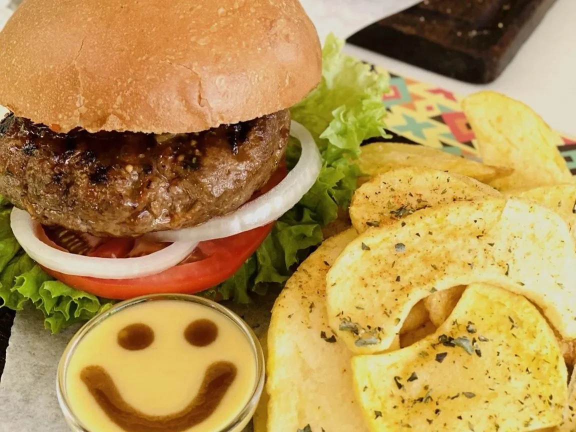 Kozis burger with lettuce, tomato, onions, and a bun, served with potato chips and a small container of yellow dipping sauce with a smiley face design.