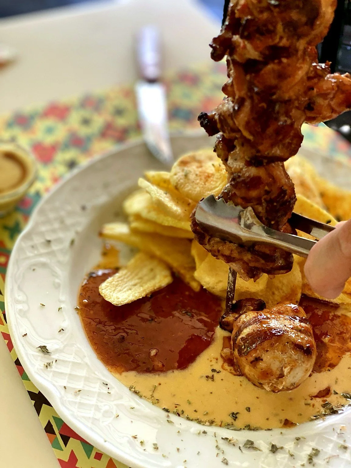 Fried chicken being dipped into a pool of sauce, with potato chips and dipping sauce on a white plate.