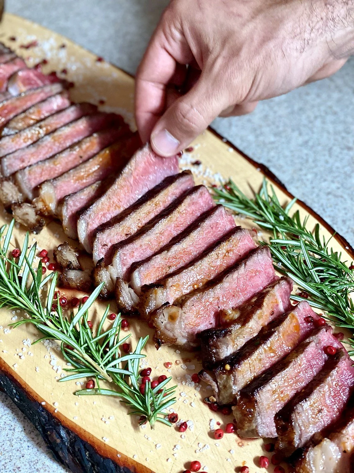 Sliced medium-rare steak on a wooden cutting board garnished with rosemary, pink peppercorns, and coarse salt.