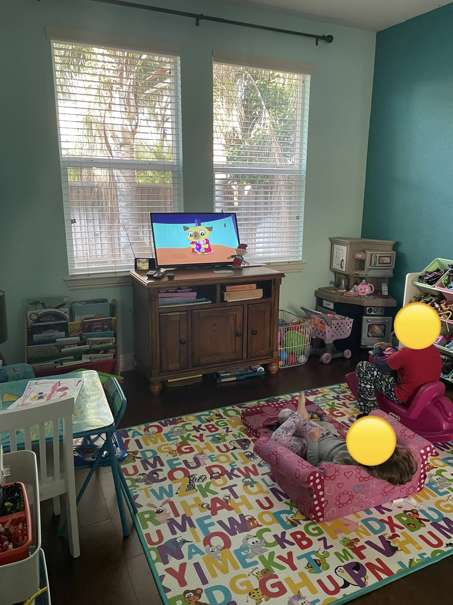 A playroom with light blue walls and large windows with blinds, showing trees outside. There is a colorful foam alphabet rug on the floor, with children playing in baby seats, one lying on her stomach and another sitting. A TV on a wooden stand displays a cartoon character. To the right is a small beige play kitchen with toys and a pink toy toaster, and various toys, books, and storage bins are visible around the room.