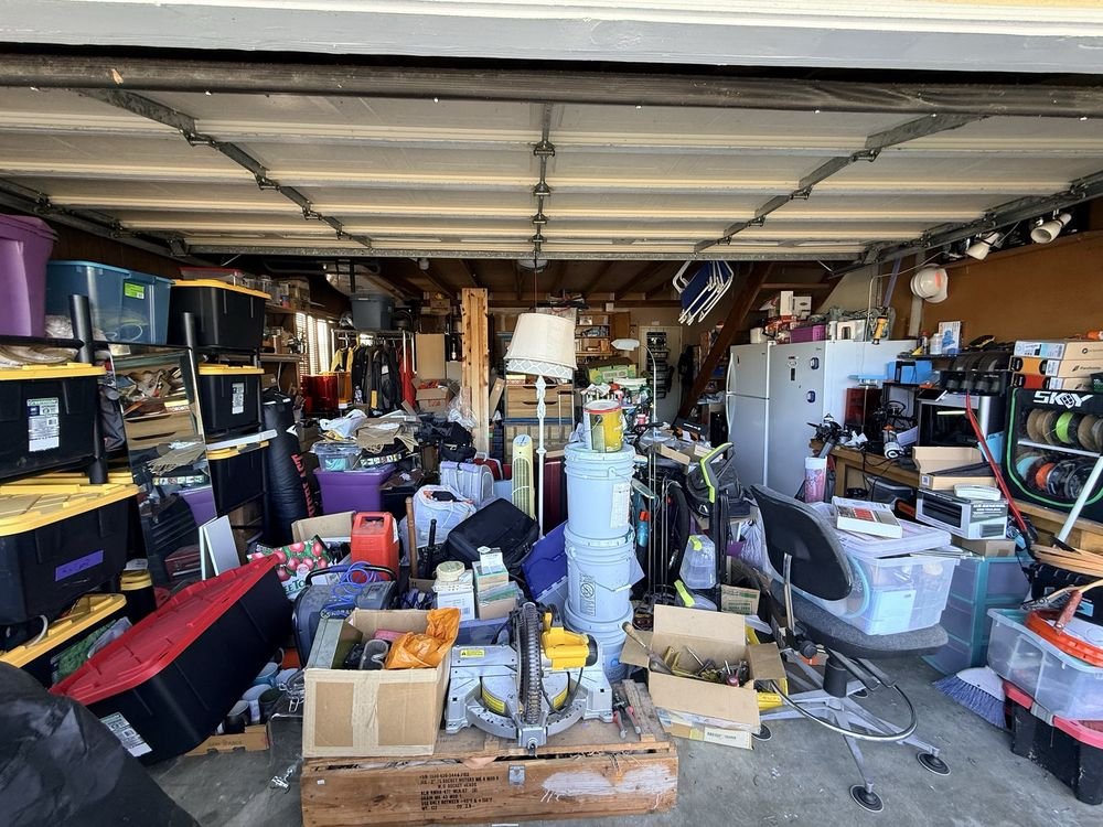 Garage filled with various storage bins, tools, and clutter, including a workbench, office chair, and ladder.