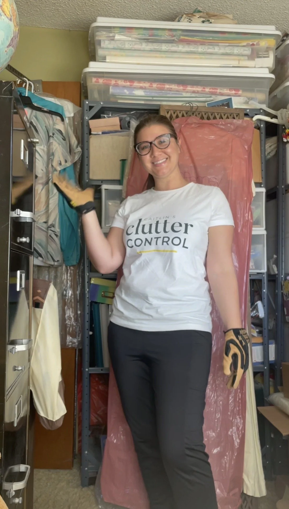 Smiling woman wearing glasses, gloves, and a white t-shirt with the words 'Caitlin's Clutter Control' standing in a cluttered storage room.