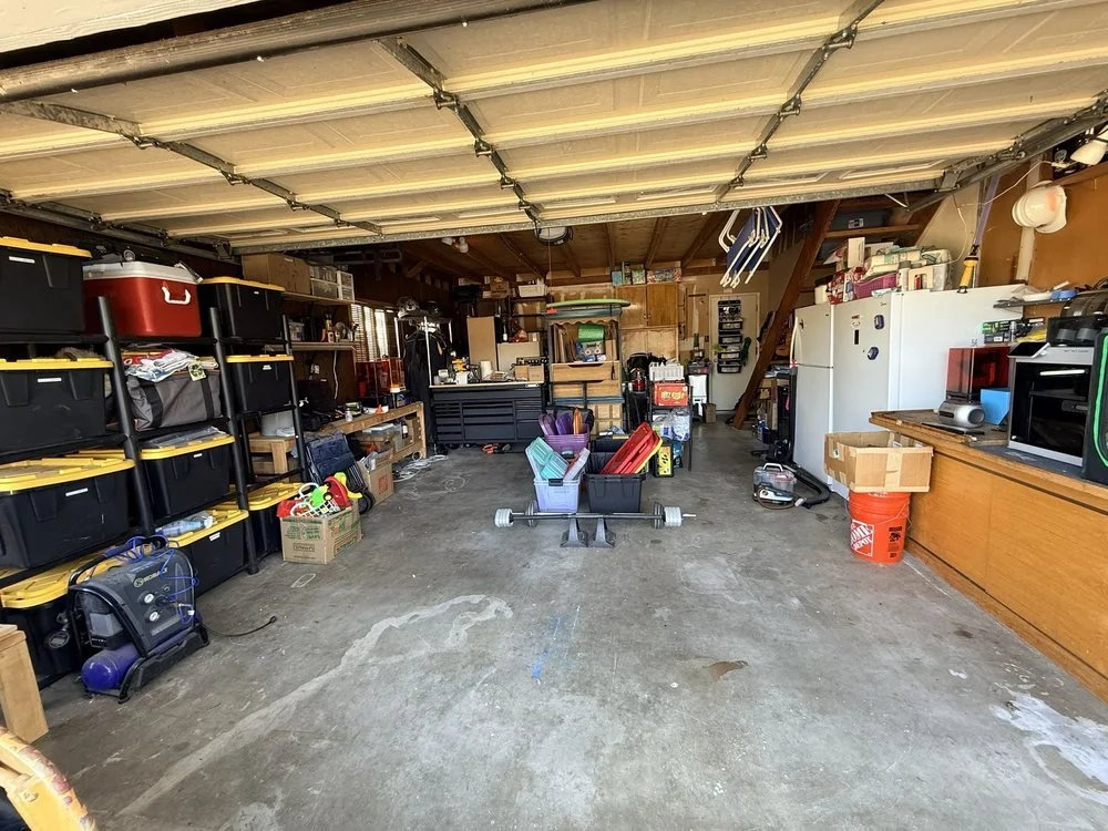 A cluttered garage with plastic storage bins, a refrigerator, tools, boxes, and various stored items.