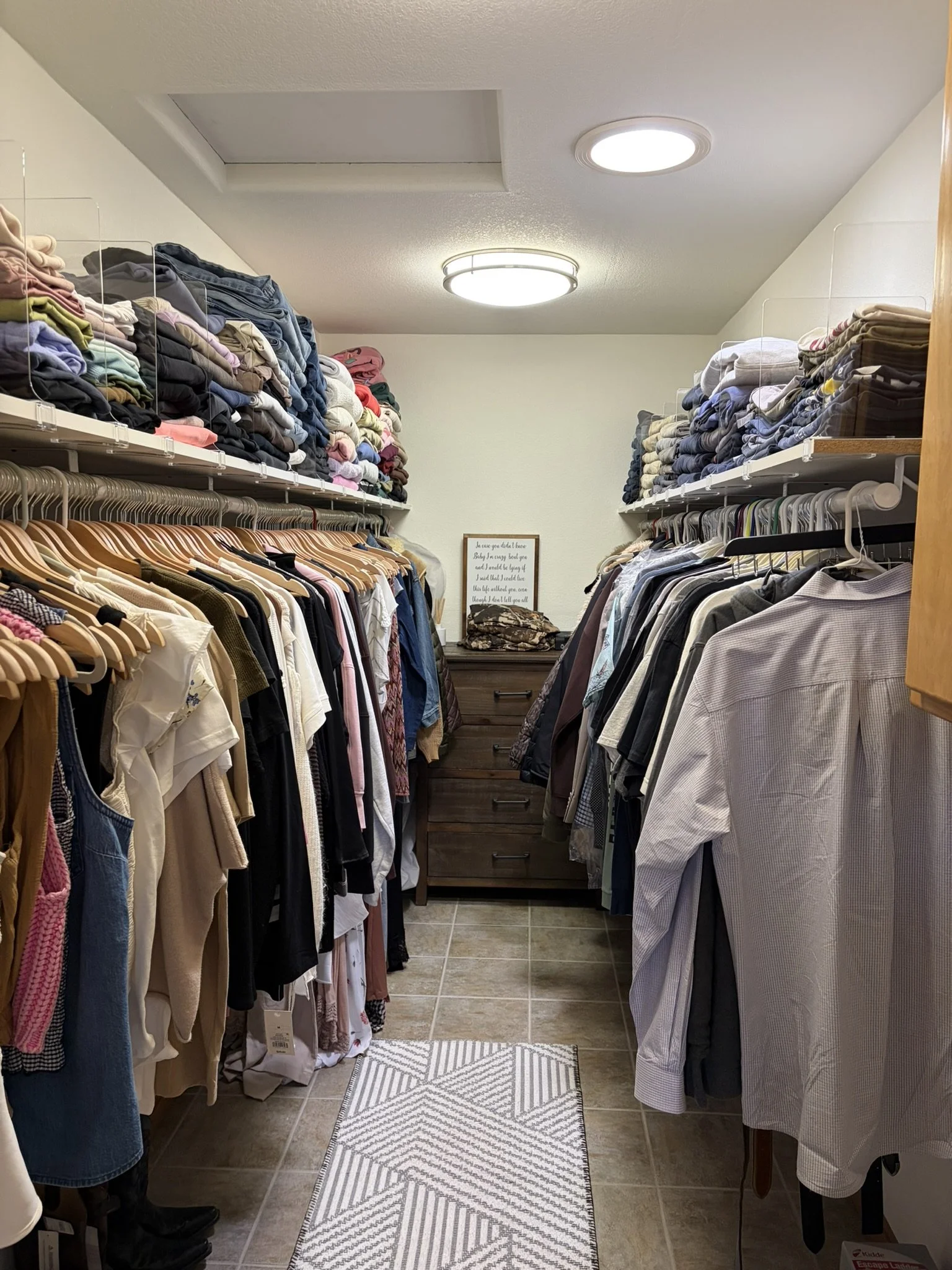 A walk-in closet with hanging clothes on both sides, a small rug in the center, and a sign on a chest of drawers at the end of the room.