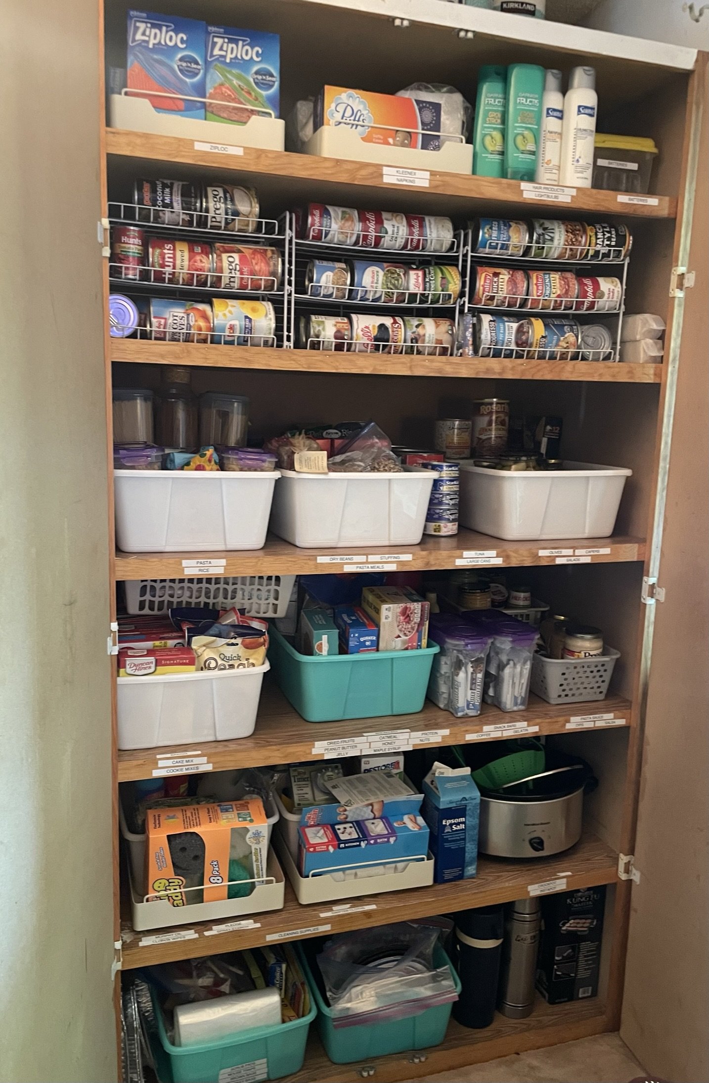 A wooden shelf filled with various food and household items, including canned goods, plastic containers, and boxes, with labeled sections for organization.