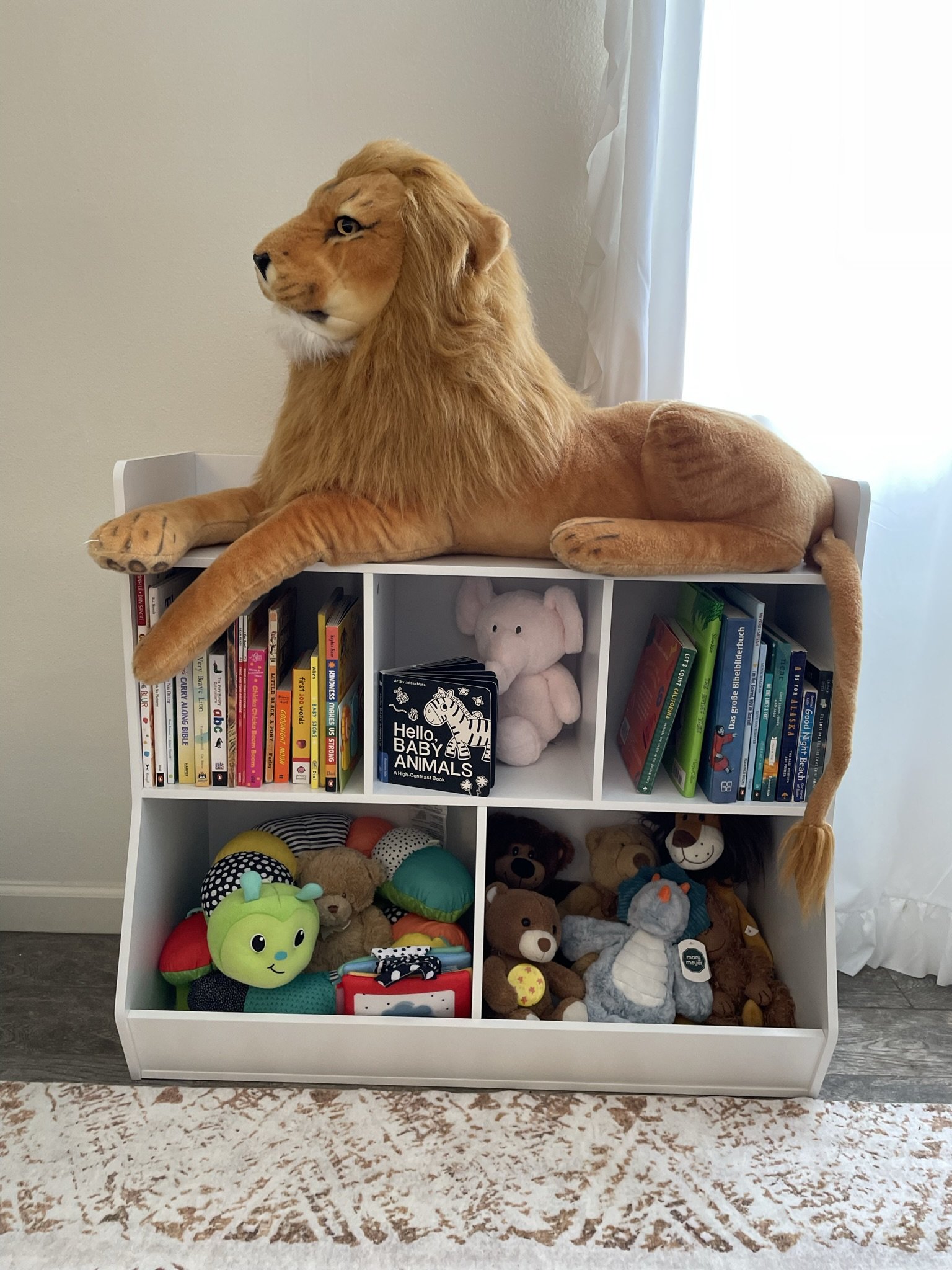 A white bookshelf topped with a plush lion that has a golden mane and tail, lying on top of it. The bookshelf contains children's books, plush toys, and stuffed animals, with a green plush fruit toy in the lower left section and a pink plush pig in the middle. The background has a plain white wall and a white curtain next to a window.