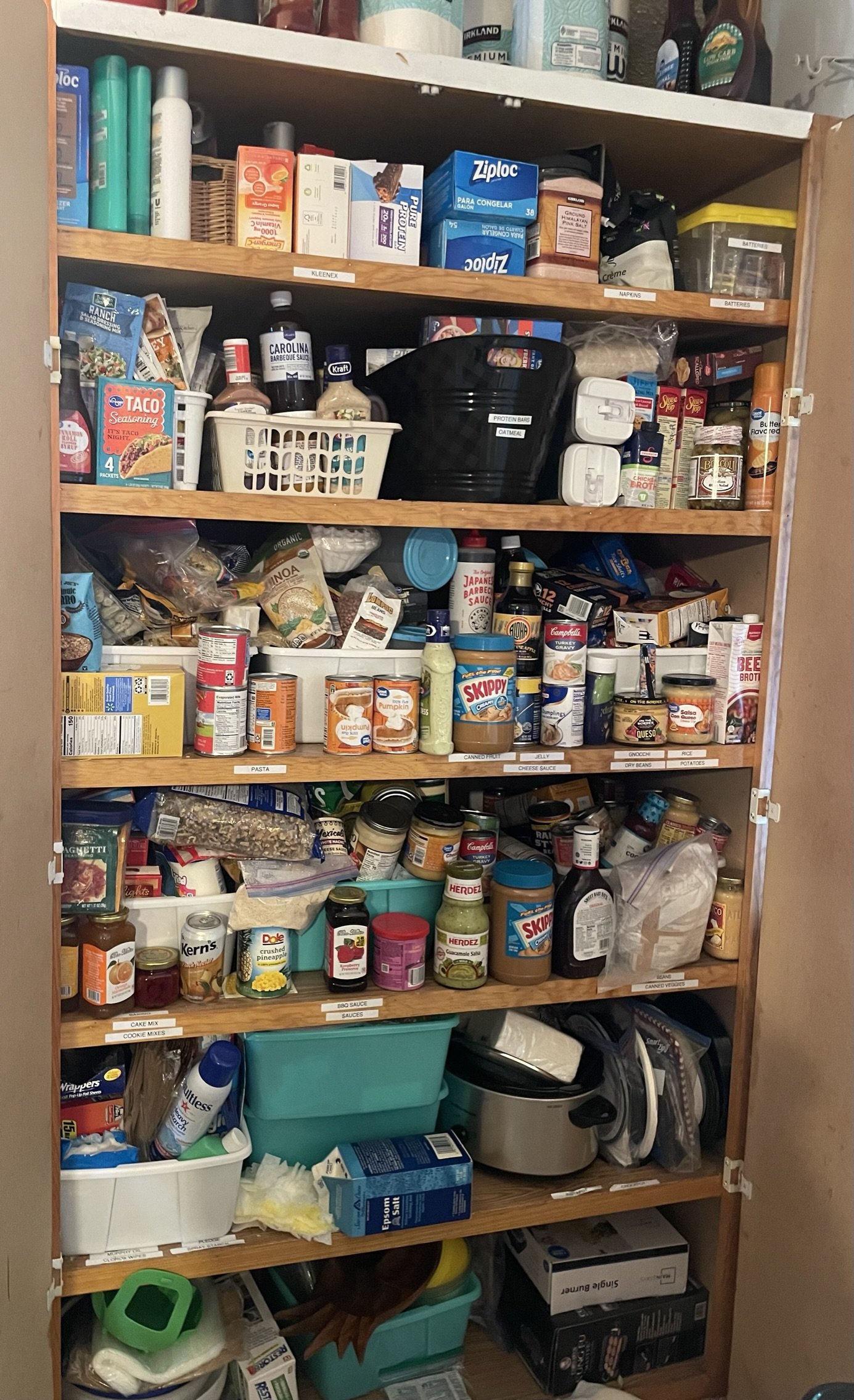 A wooden pantry shelf filled with various food products, condiments, canned goods, snacks, and household items organized in plastic containers and on labeled shelves.