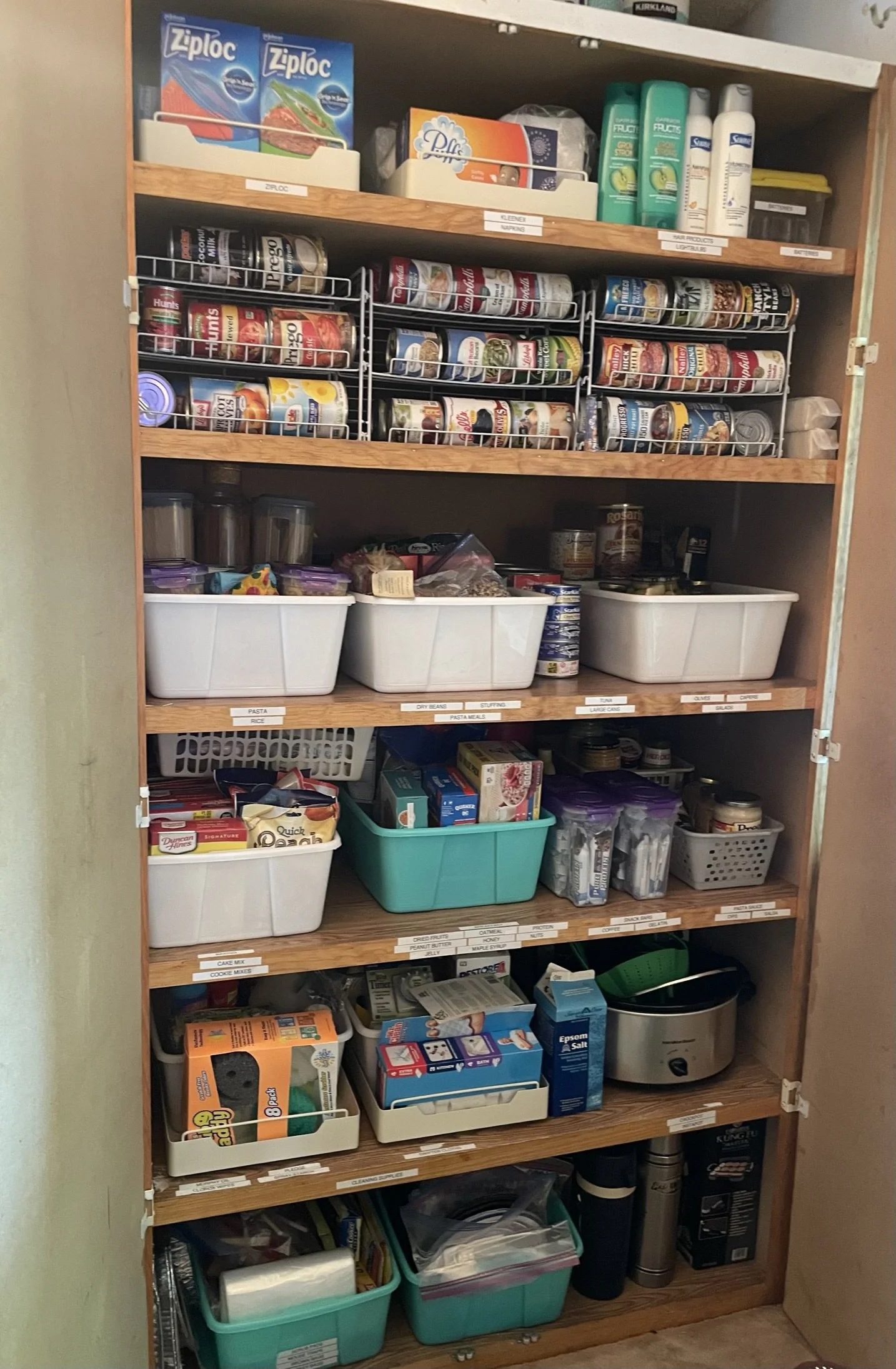 A tall wooden shelf filled with various organized household items including canned foods, plastic containers, cleaning supplies, and miscellaneous kitchen supplies. The shelf has labeled sections for easier organization.