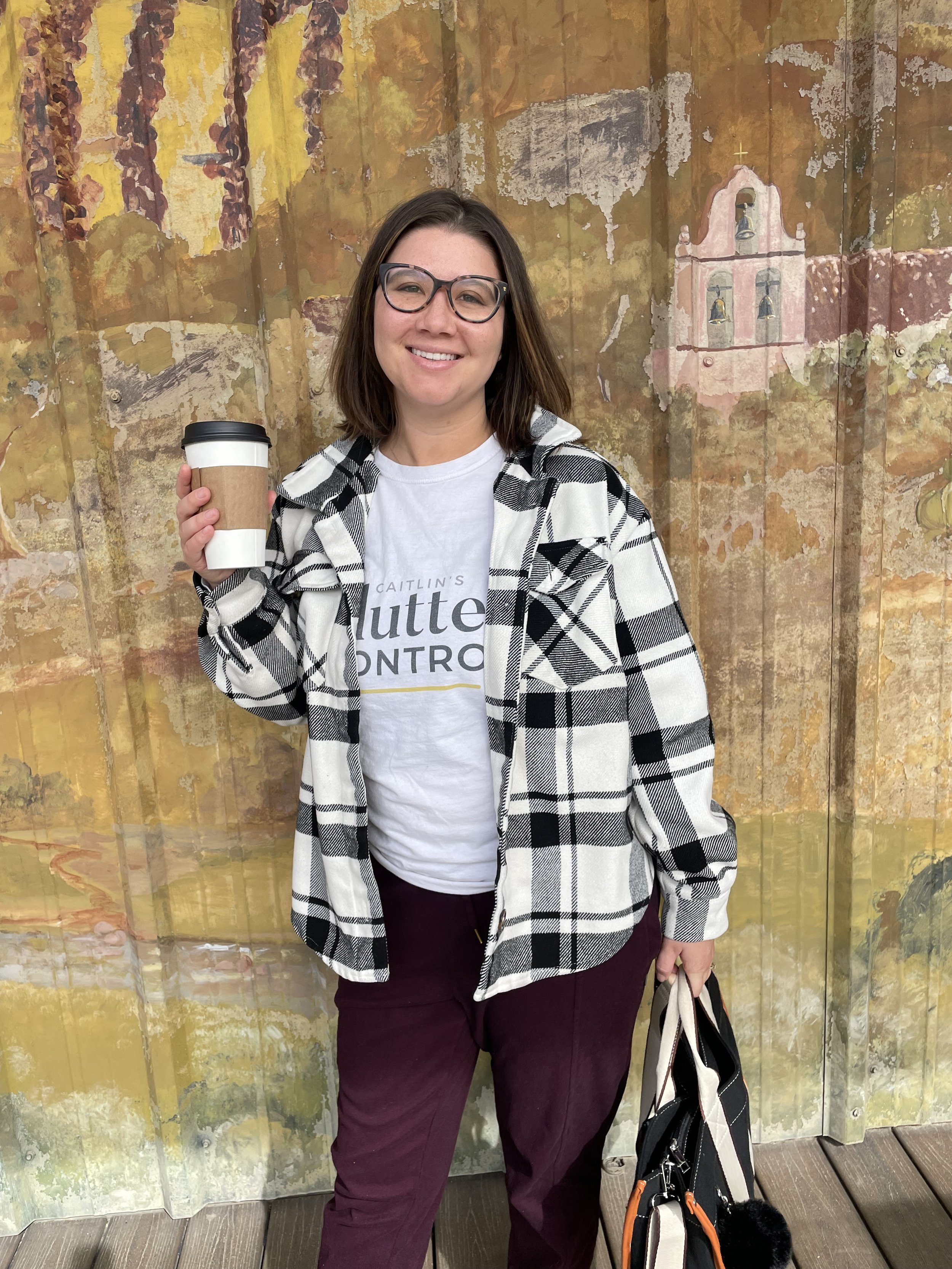 A woman with shoulder-length brown hair, glasses, and a white T-shirt under a black and white plaid shirt is smiling and holding a takeaway coffee cup. She is standing in front of a weathered, painted wall with a church illustration in the background. She has maroon pants and is holding a tote bag in her left hand, with a backpack on the ground beside her.