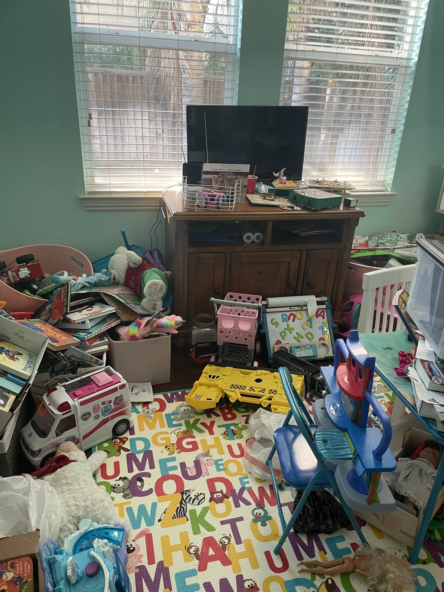 A cluttered room with a TV on a wooden cabinet, toys, books, a colorful alphabet mat, a pink toy shopping cart, and various children’s items scattered around, near two large windows with blinds.