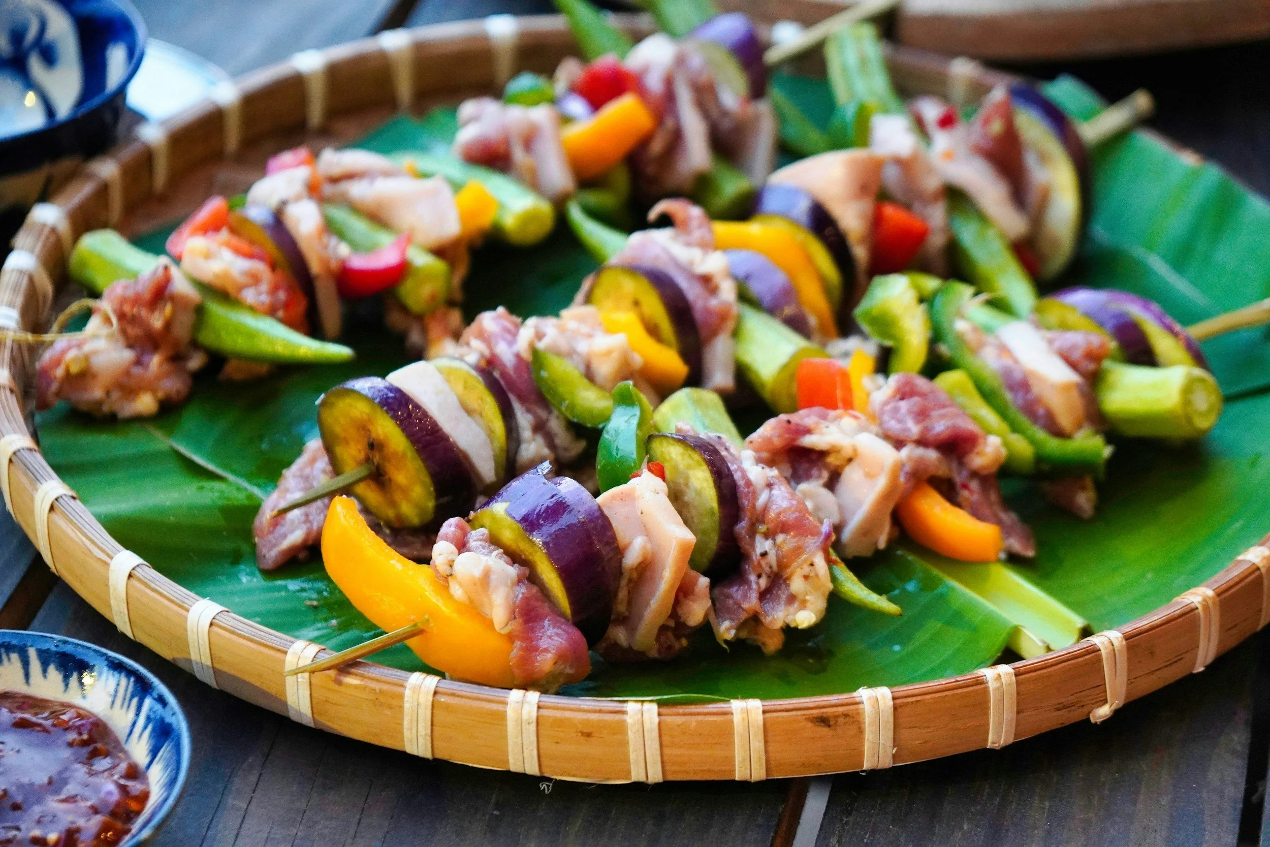 Skewers with grilled vegetables and meat on a green banana leaf served in a round bamboo tray.