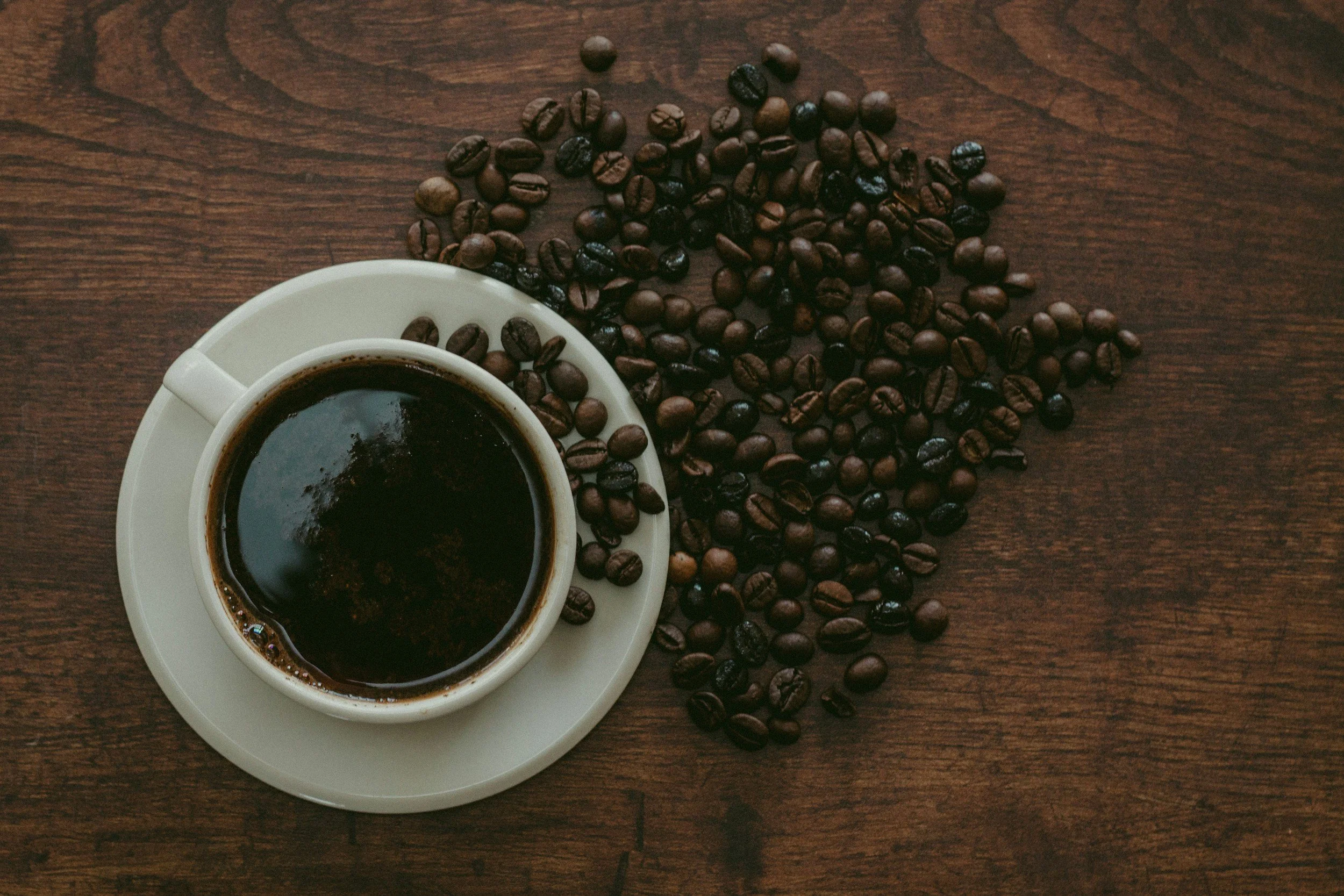 A cup of black coffee on a saucer surrounded by scattered coffee beans on a wooden table.