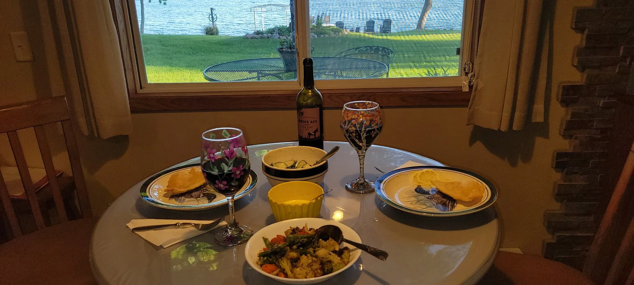 A cozy indoor dining table set for a meal with two plates of food, a bowl of pickles, a bowl of mixed vegetables, and a glass of wine. There is a bottle of wine labeled 'Broke Ass' on the table. The table is near a window showing a lakeside view with