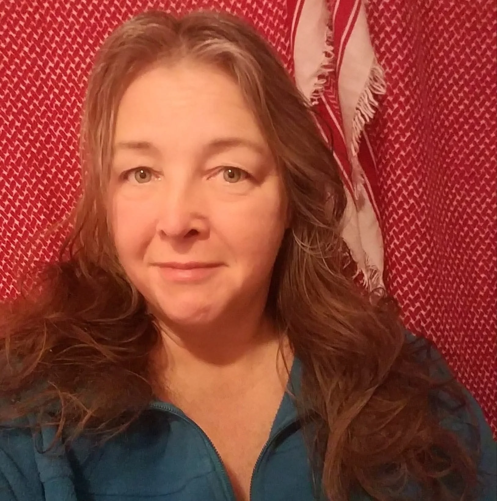 A woman with light brown, curly hair and light skin is lying on a red patterned blanket with fringe, smiling slightly and looking at the camera.