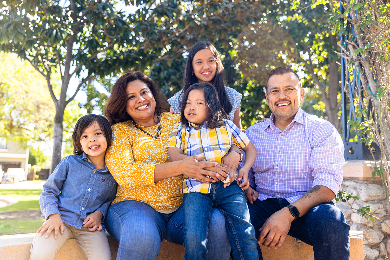 A multigenerational family of six sitting together outdoors on a sunny day, smiling, with trees and greenery in the background.