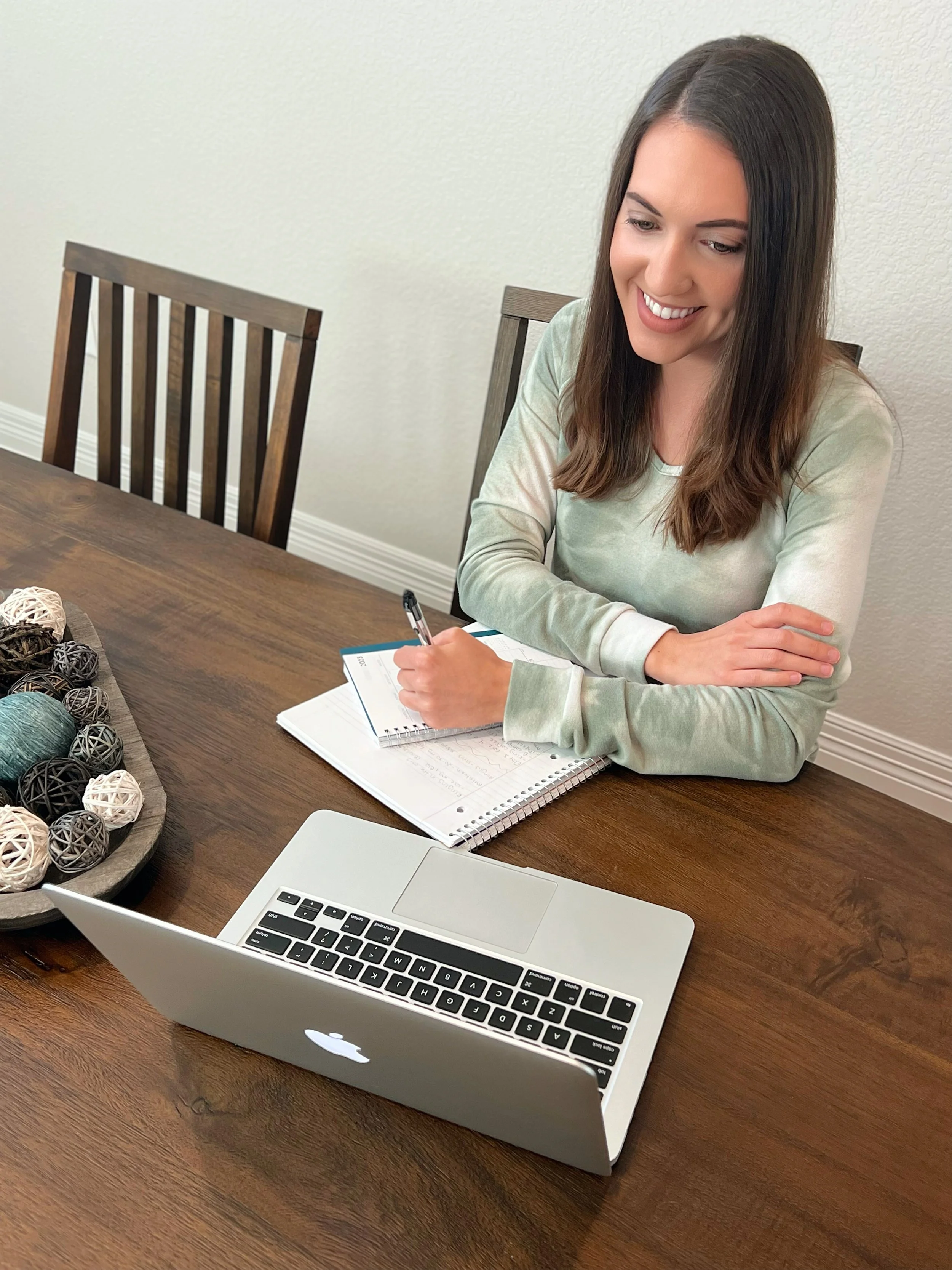 A woman sitting at a wooden table with a MacBook laptop, a spiral notebook, and a pen. She is smiling and looking down, with a decorative tray of yarn balls to her left.