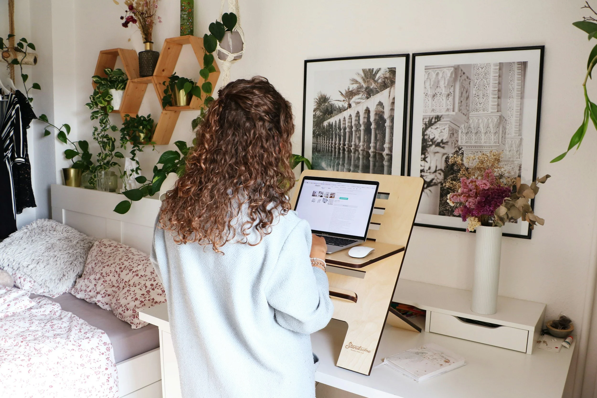 A person with curly hair working on a laptop at a desk in a cozy, decorated room with green plants, framed pictures, and floral pillows.