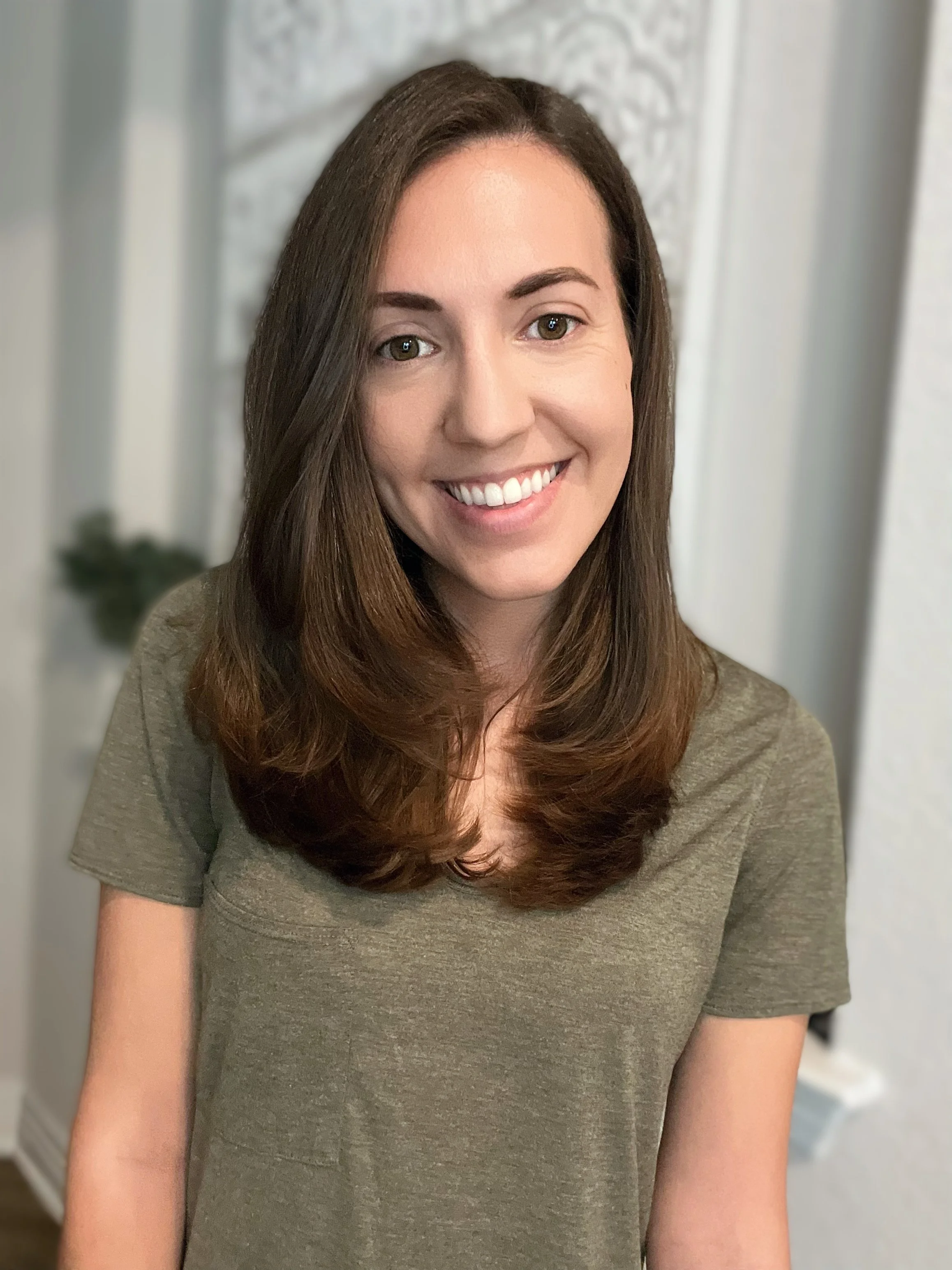 A woman with long brown hair smiling while wearing a gray T-shirt. The background shows a decorative white wall and part of a green plant.