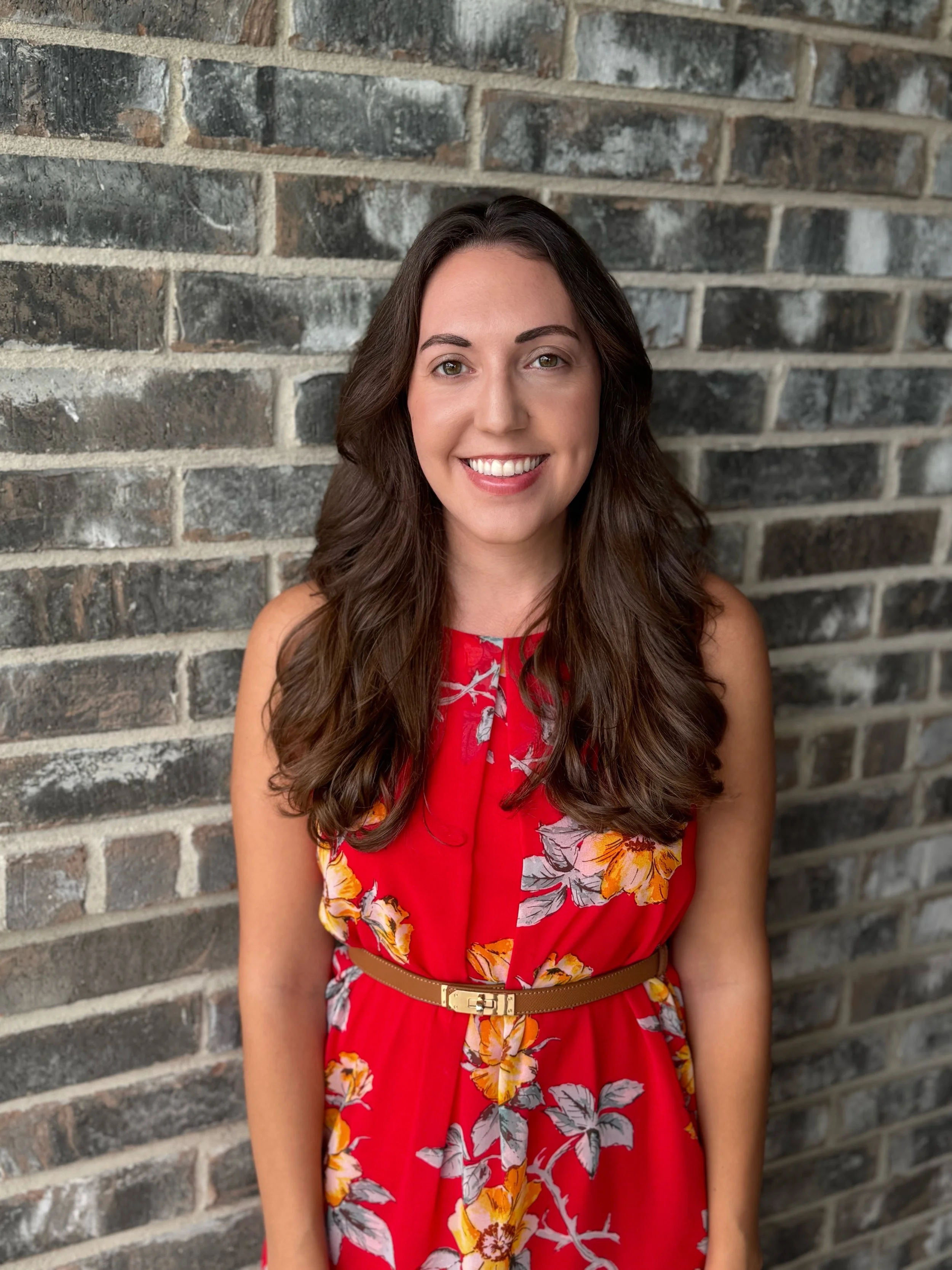 A woman with long, wavy brown hair and green eyes, smiling and wearing a red sleeveless dress with yellow, pink, and white floral pattern, standing in front of a gray brick wall.