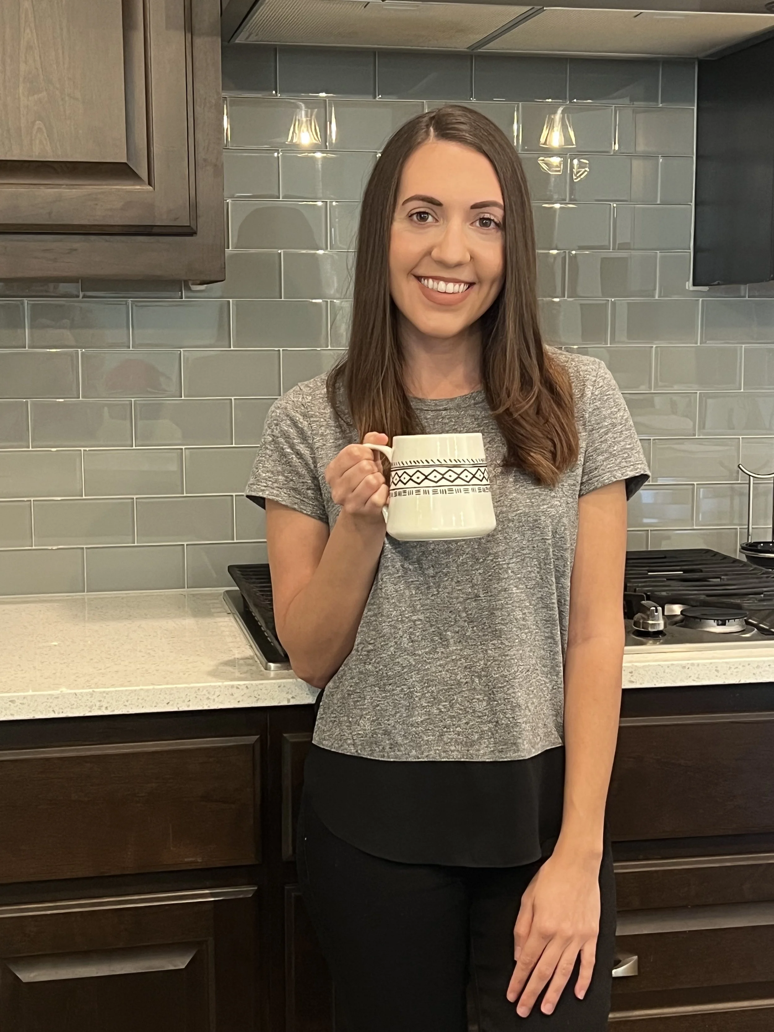 A woman with brown hair holding a mug in a kitchen with gray subway tile backsplash, dark wood cabinets, and a stovetop.