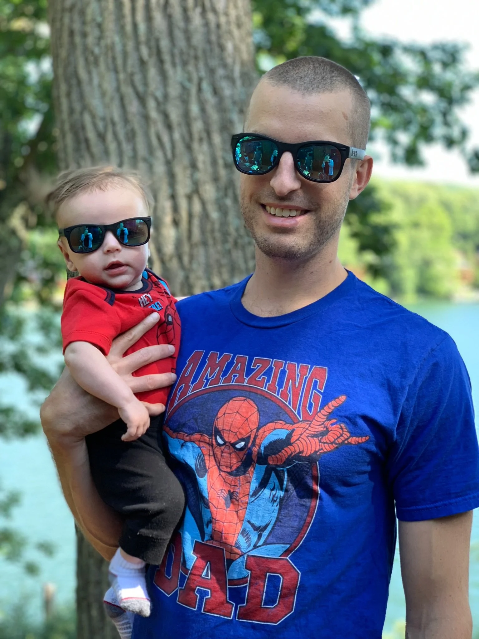 A man holding a young child outdoors with a large tree trunk and green foliage in the background. Both are wearing sunglasses and casual clothing.