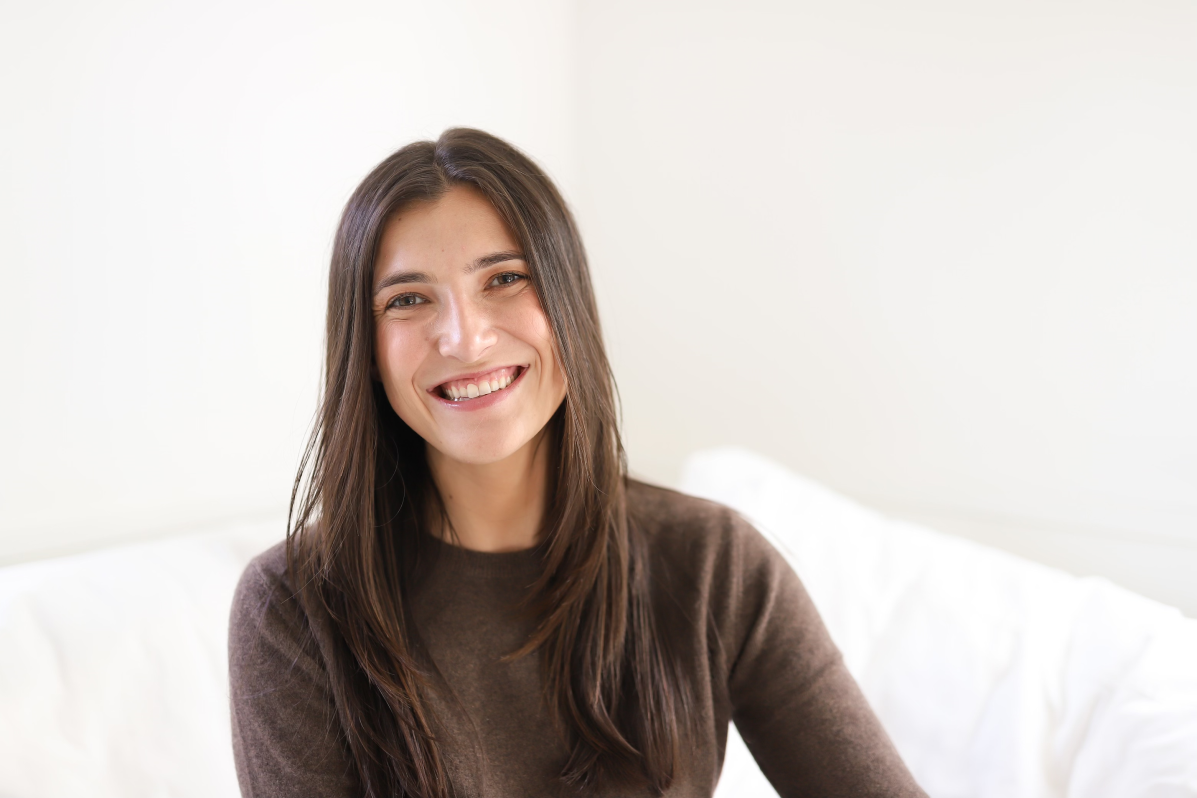 A young woman with long brown hair smiling and looking at the camera, sitting in a bright room with a white wall background.
