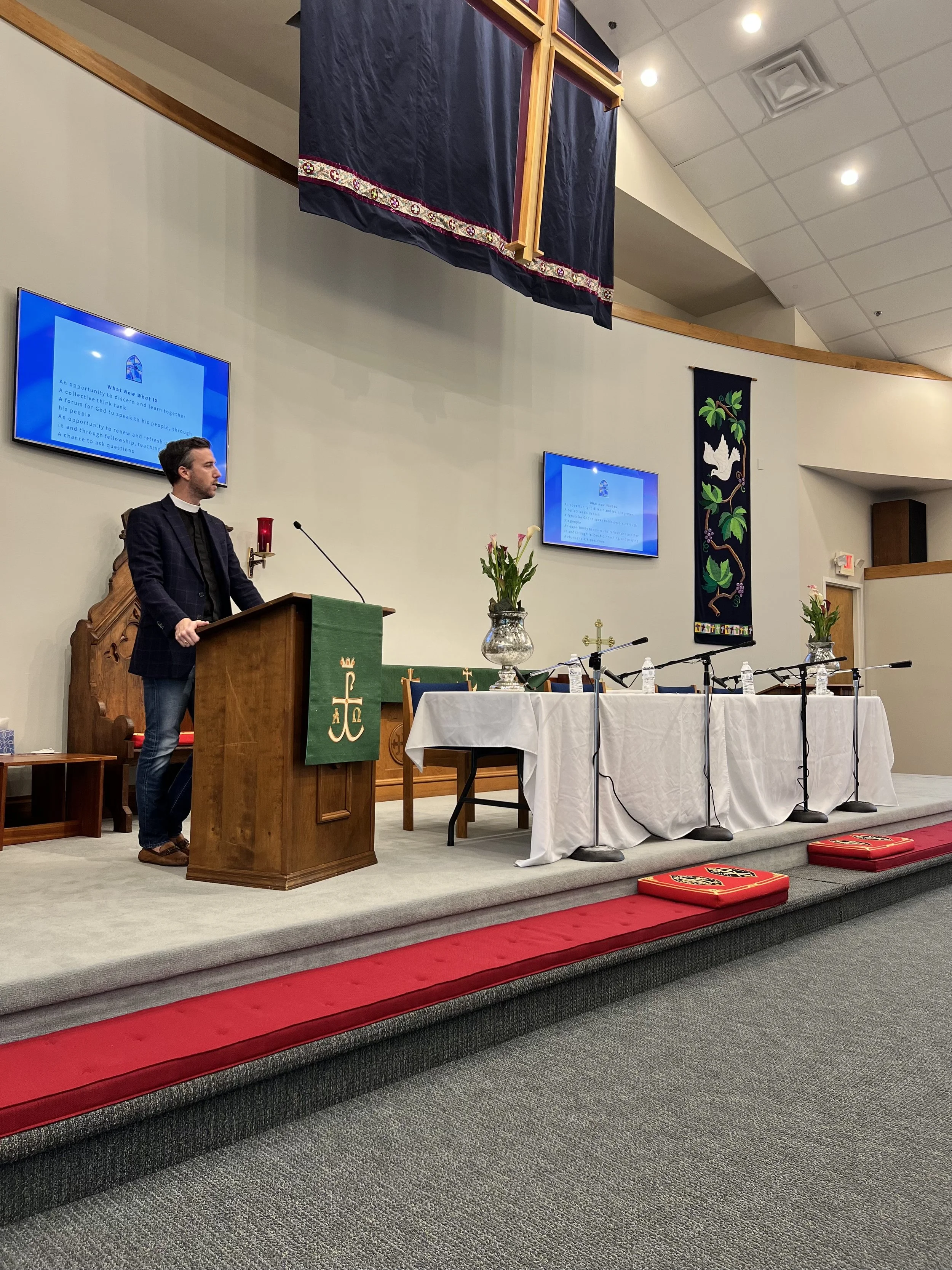 A man wearing a dark blazer and a clerical collar is standing at a wooden pulpit in a church, with a microphone. Behind him is a large screen displaying a PowerPoint slide and a green banner with gold LDS symbols. The altar is covered with a white cloth and decorated with flowers and water bottles. The background includes religious banners, a hanging cross, and a red carpet on the stage. The ceiling has white tiles and ceiling lights.