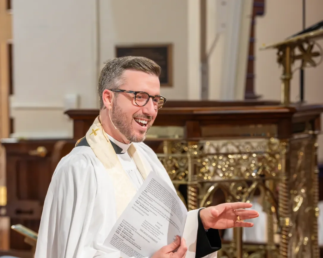 A smiling man wearing glasses, a white clerical robe, and a stole with gold crosses, holding a booklet, standing in front of an ornate church altar.