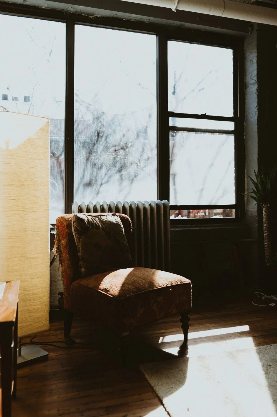 A vintage armchair with floral upholstery positioned near a large window with snow outside, a radiator behind the chair, and a potted plant in the corner.