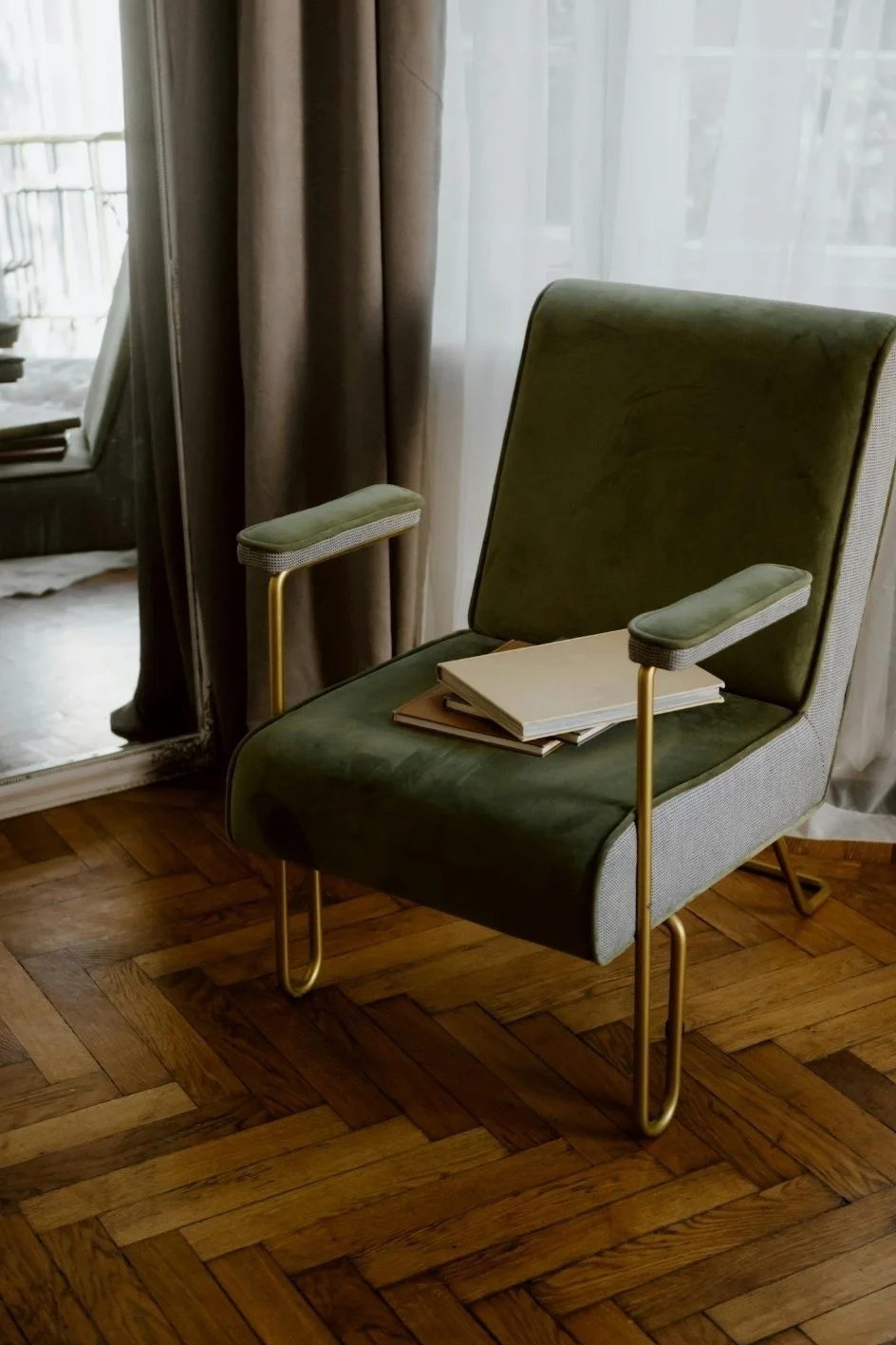A green armchair with gold metal armrests and legs, placed on a wooden floor near a window with white curtains. A few books are stacked on the seat.